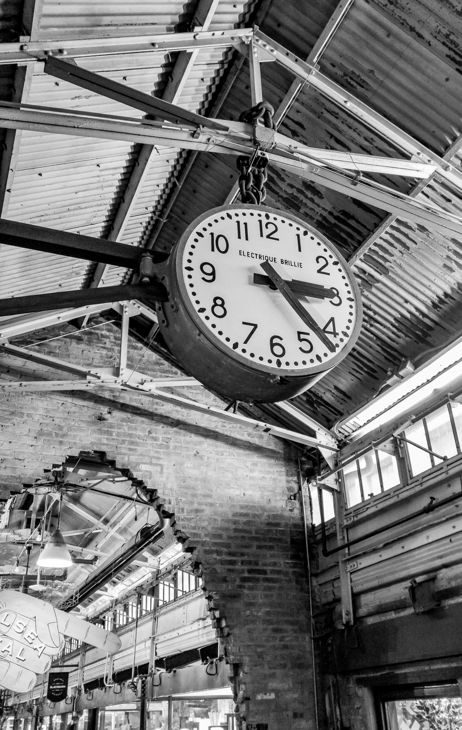 A large antique clock hangs from the rafters of a brick building with other clocks visible below.