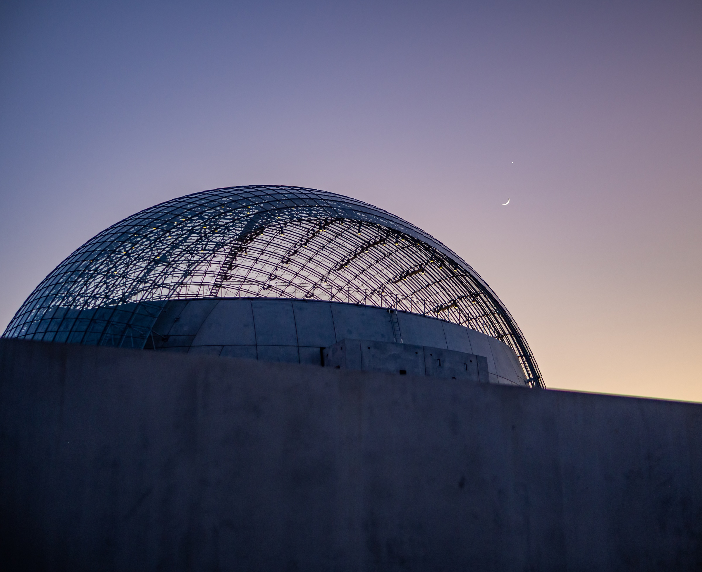 A large, modern dome building sits against a twilight sky with a faint crescent moon.