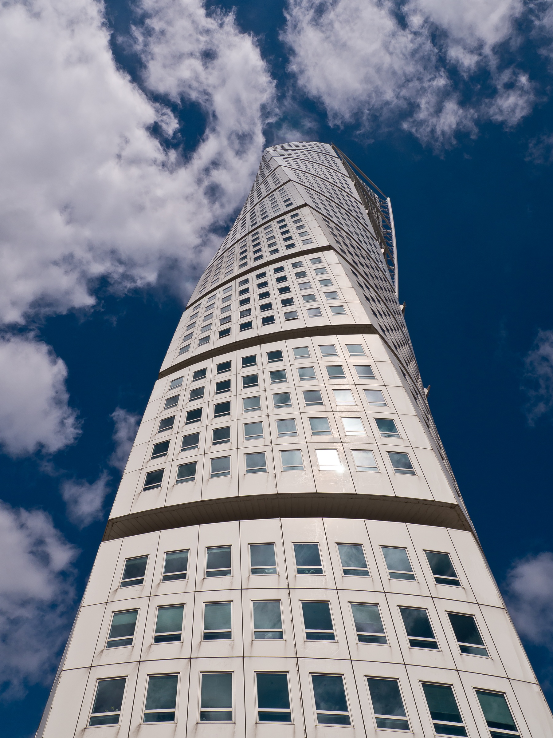 A tall, twisting white skyscraper rises against a bright blue sky with fluffy clouds.