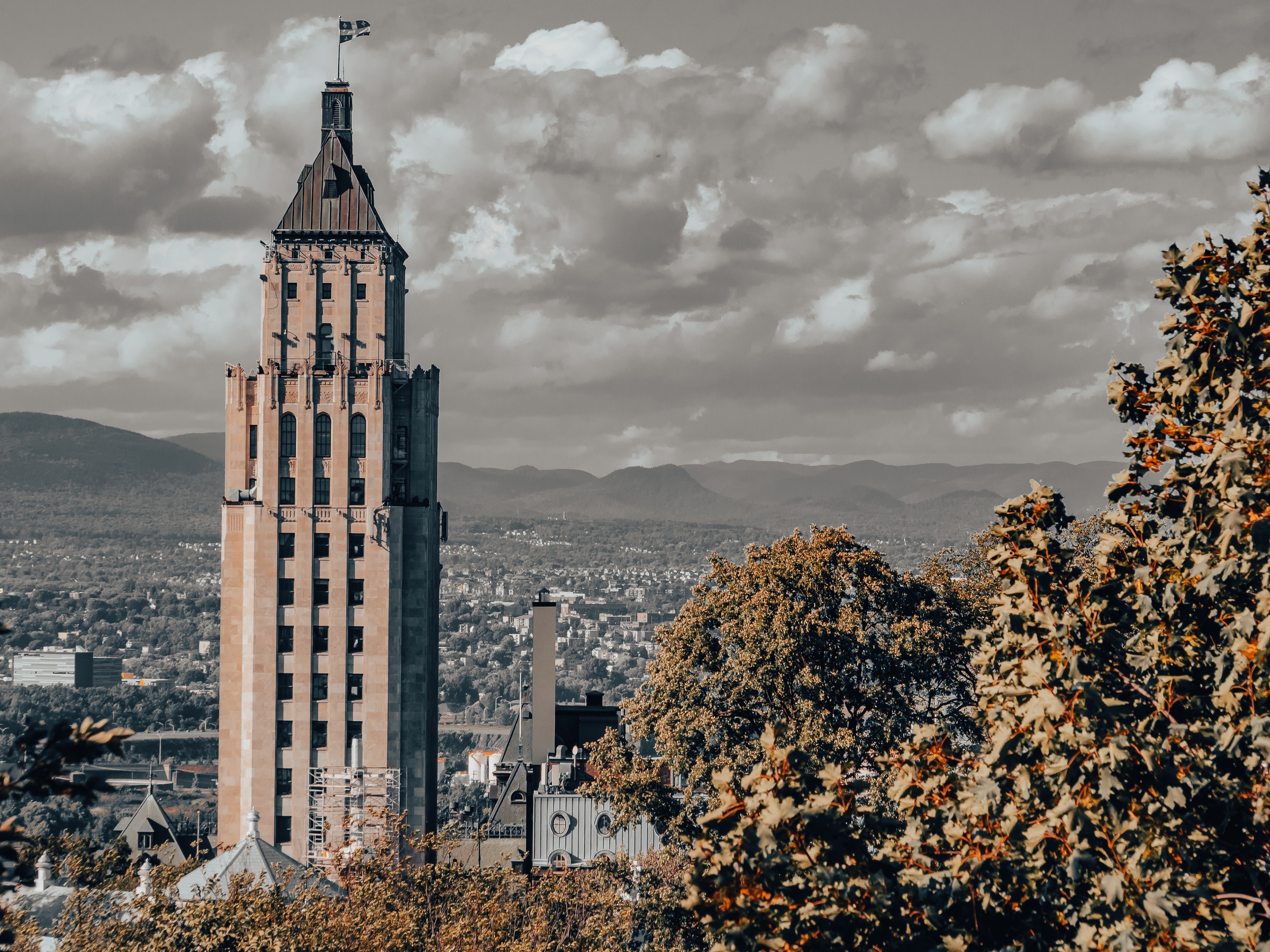 A tall tower rises above a city landscape with distant mountains under a cloudy sky.