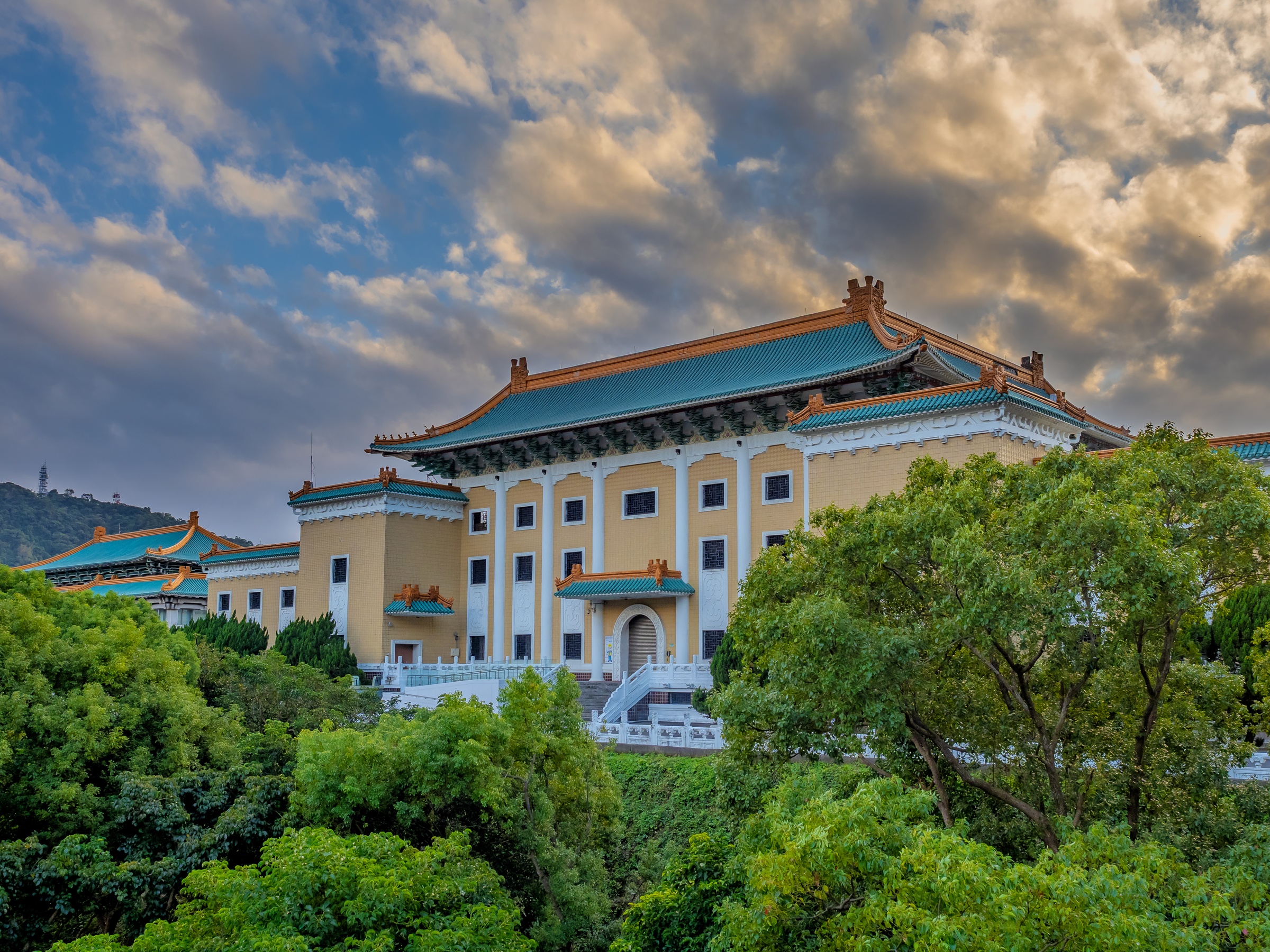 A large, ornate yellow building with a green roof sits nestled amongst lush greenery under a cloudy sky.