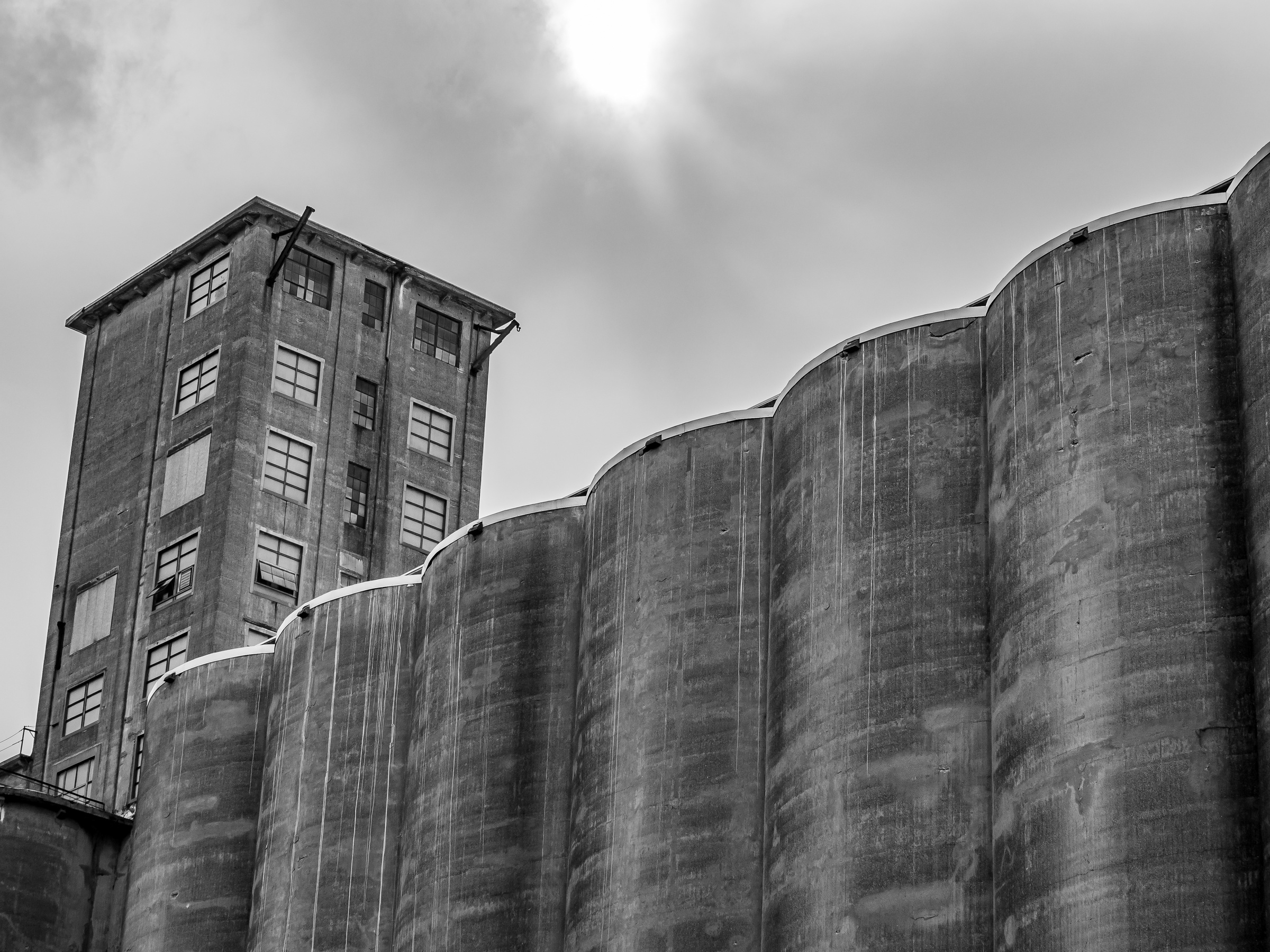 Monolithic concrete silos and a taller building rise against a cloudy sky.