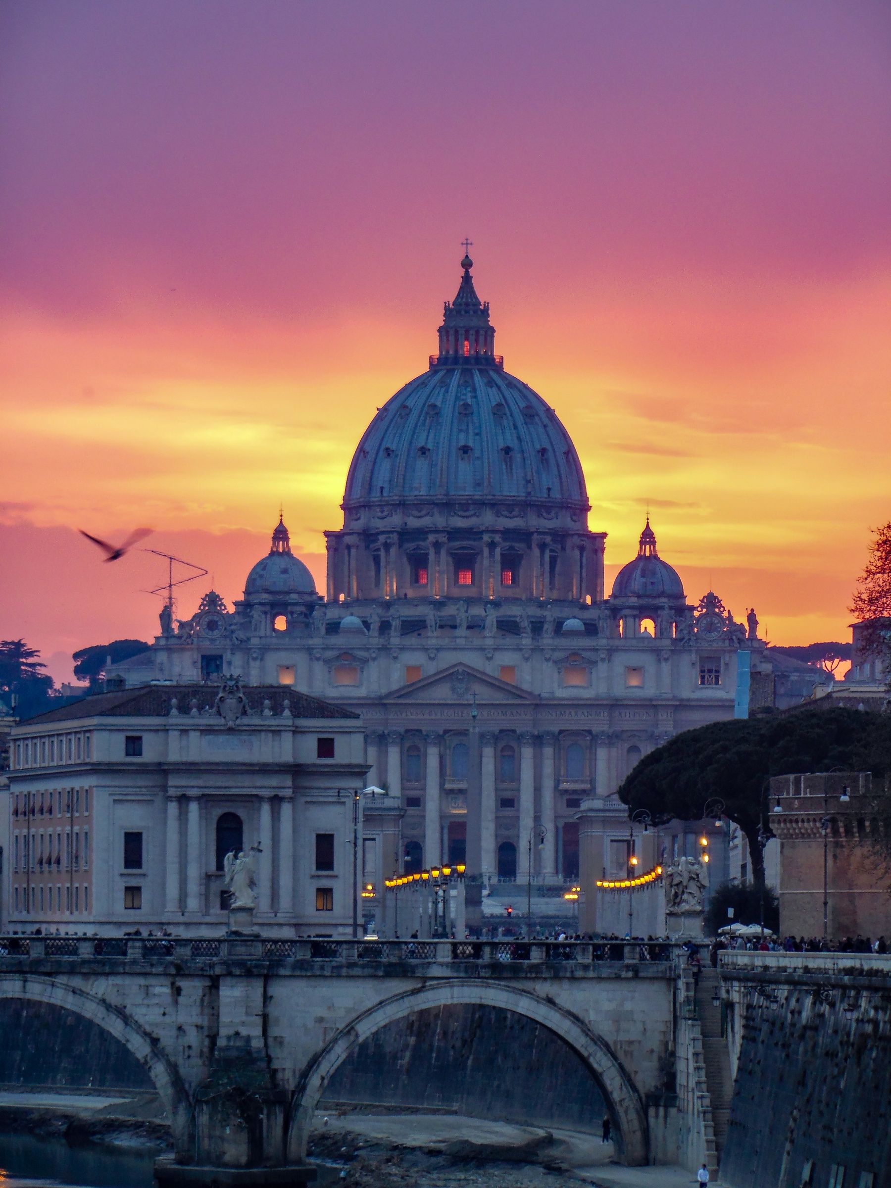 St. Peter's Basilica sits beneath a vibrant sunset sky, framed by an arched bridge and surrounding buildings.
