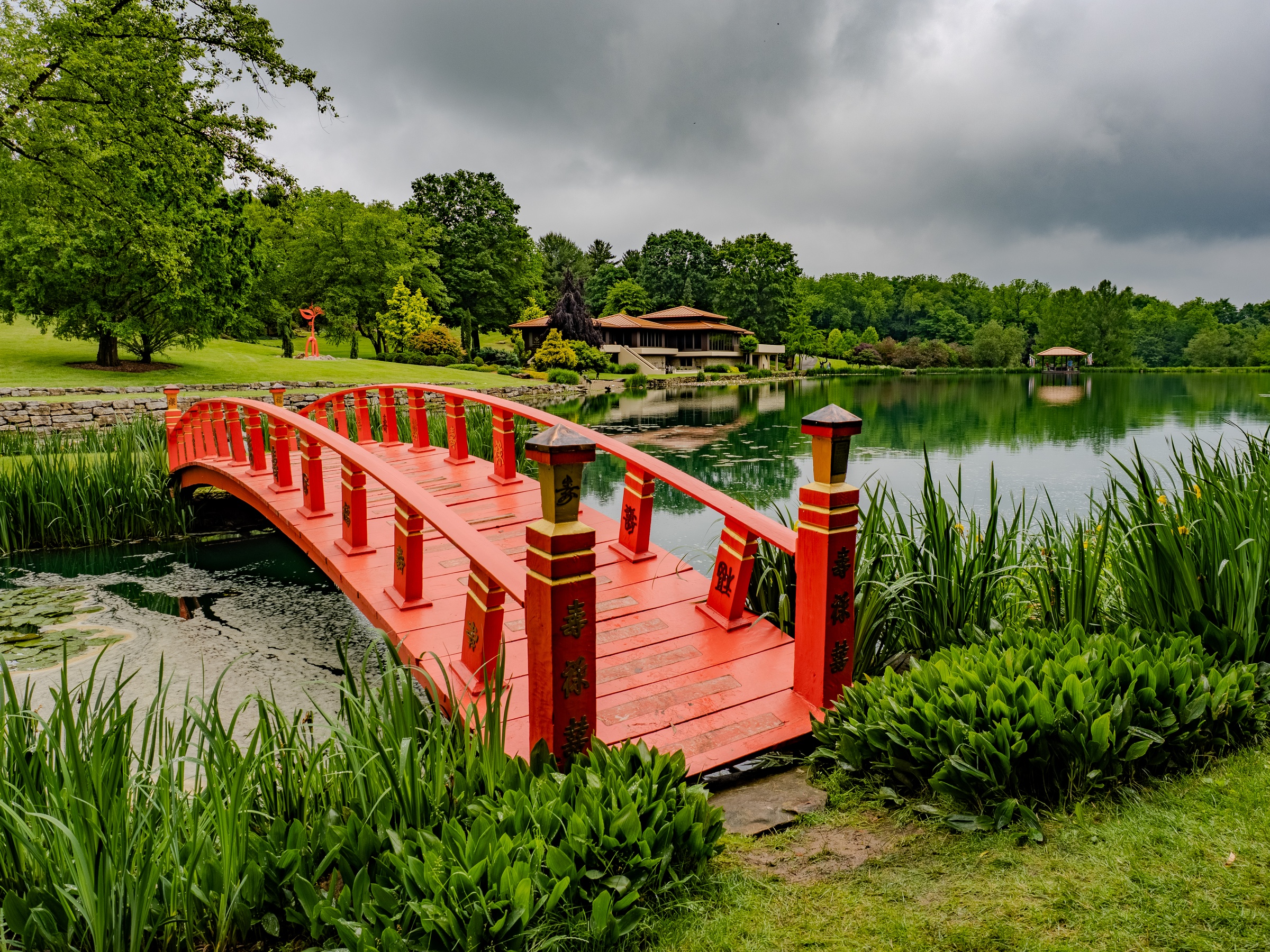 A vibrant red arched bridge spans a calm lake surrounded by lush green trees and foliage.