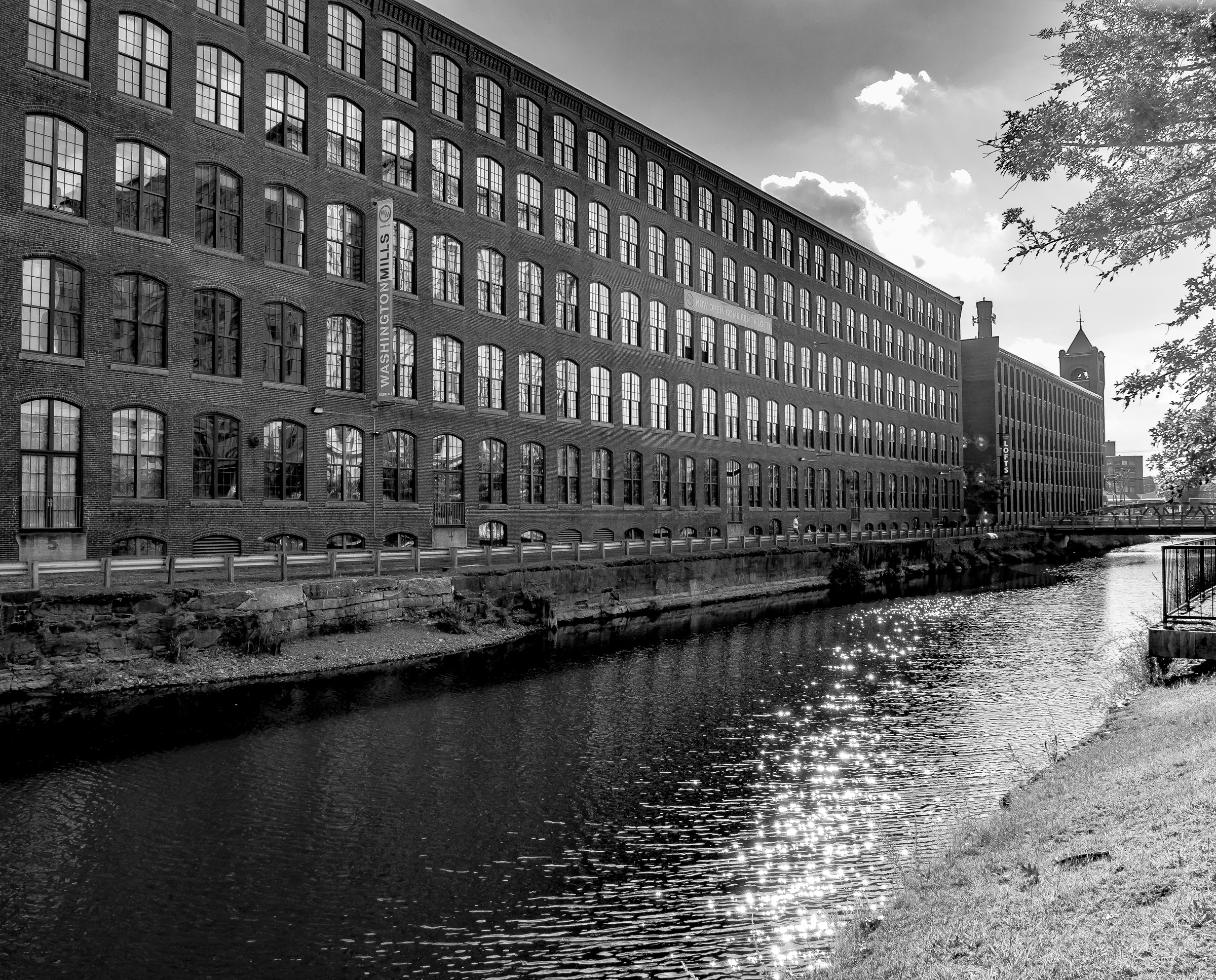 A large brick building with many windows sits alongside a river, under a cloudy sky.