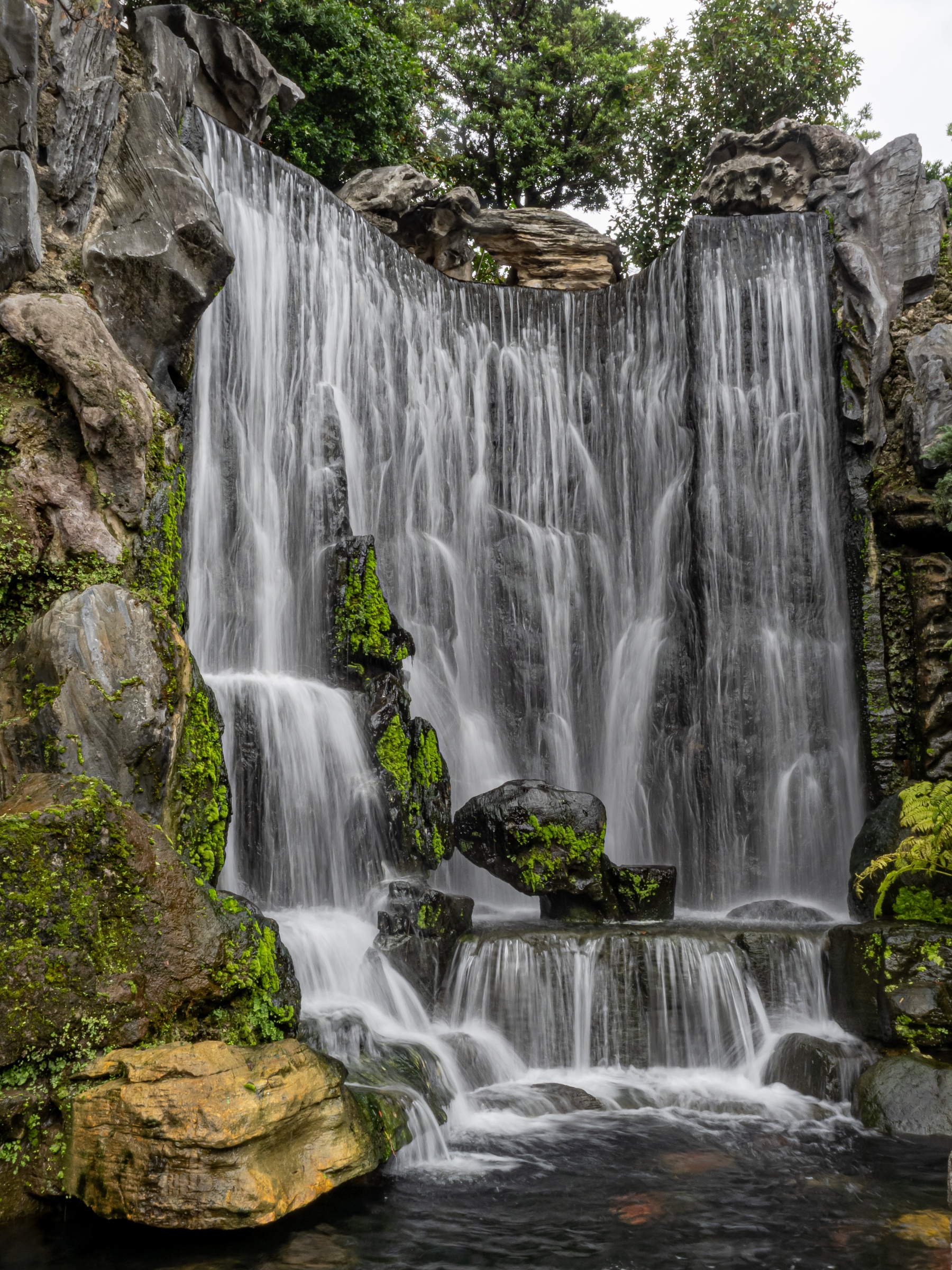 A tiered waterfall cascades down mossy rocks within a lush, landscaped garden setting.