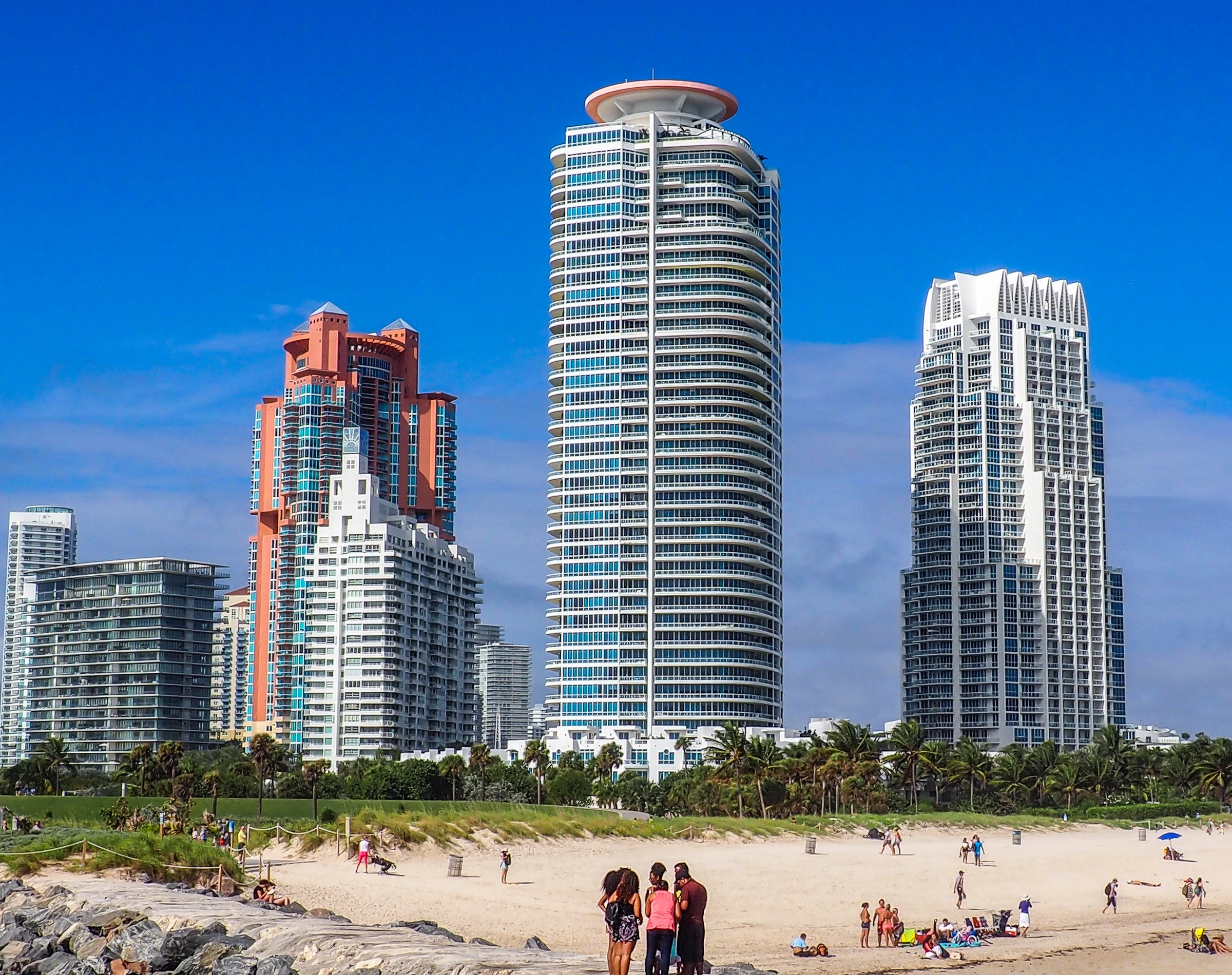 Miami's beachfront skyline features several tall, modern buildings against a bright blue sky and sandy beach.