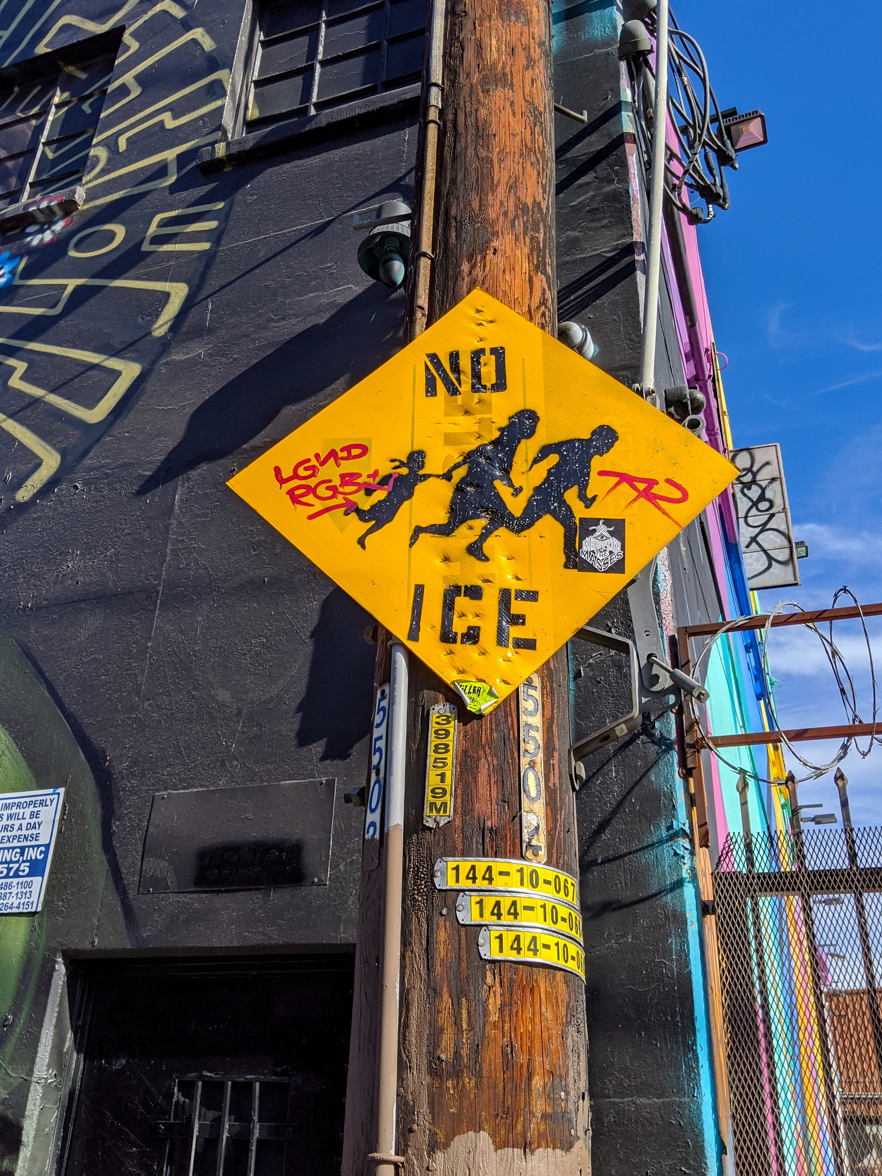 A yellow diamond sign with graffiti depicting running figures is affixed to a weathered utility pole against a building.