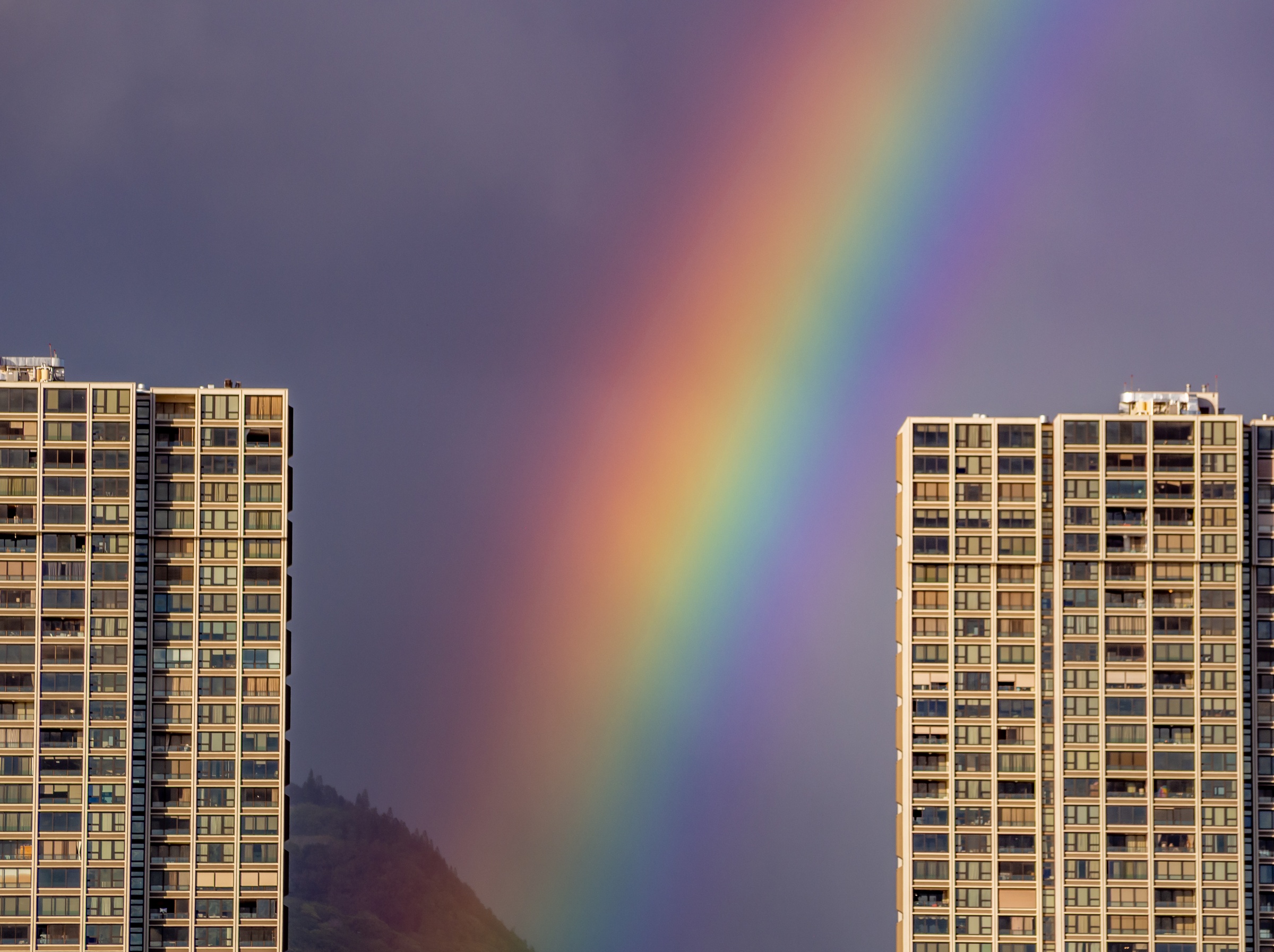 A vibrant rainbow arches between two tall apartment buildings against a cloudy sky.