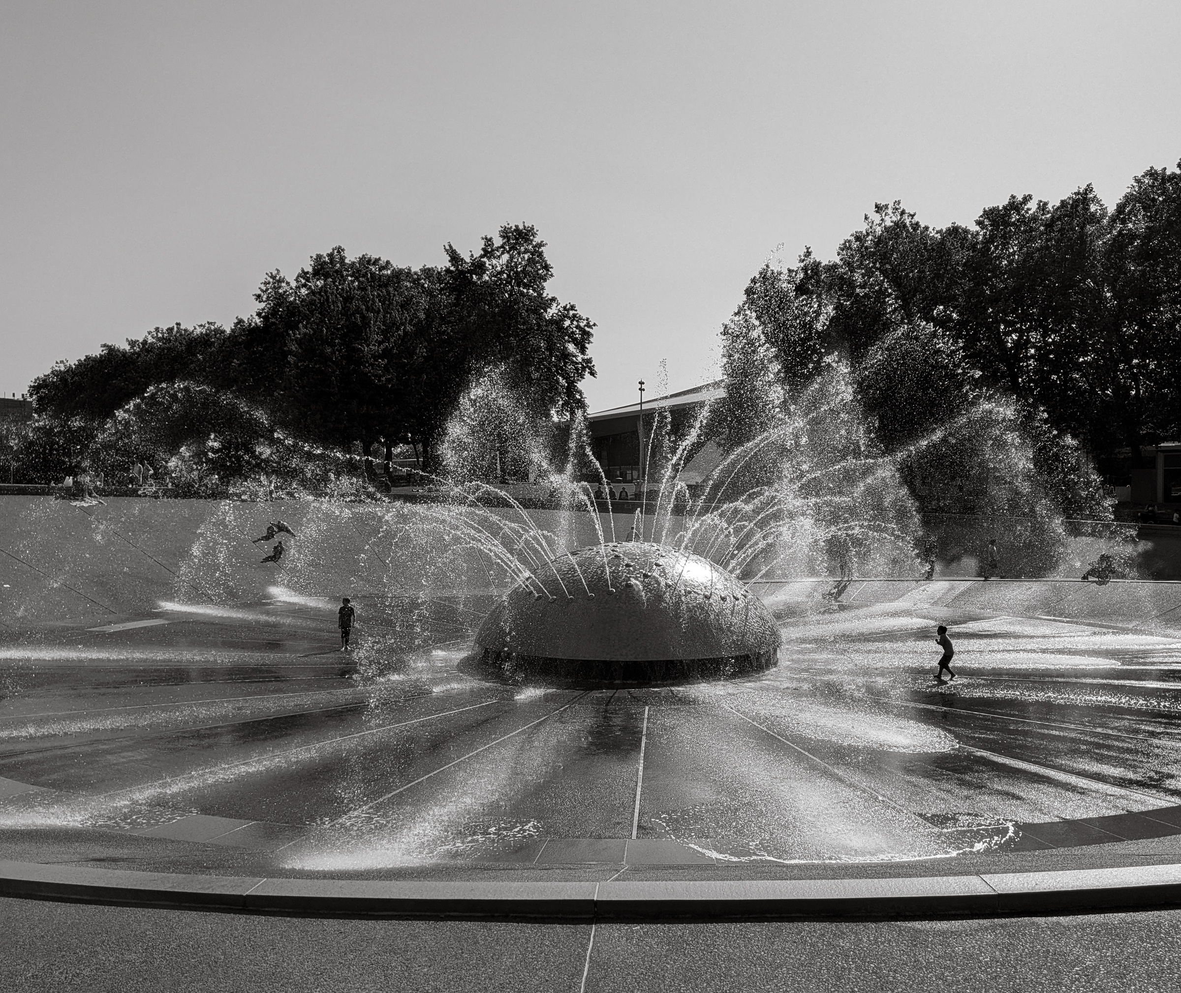 A fountain sprays water in a plaza with people playing amidst trees and buildings.