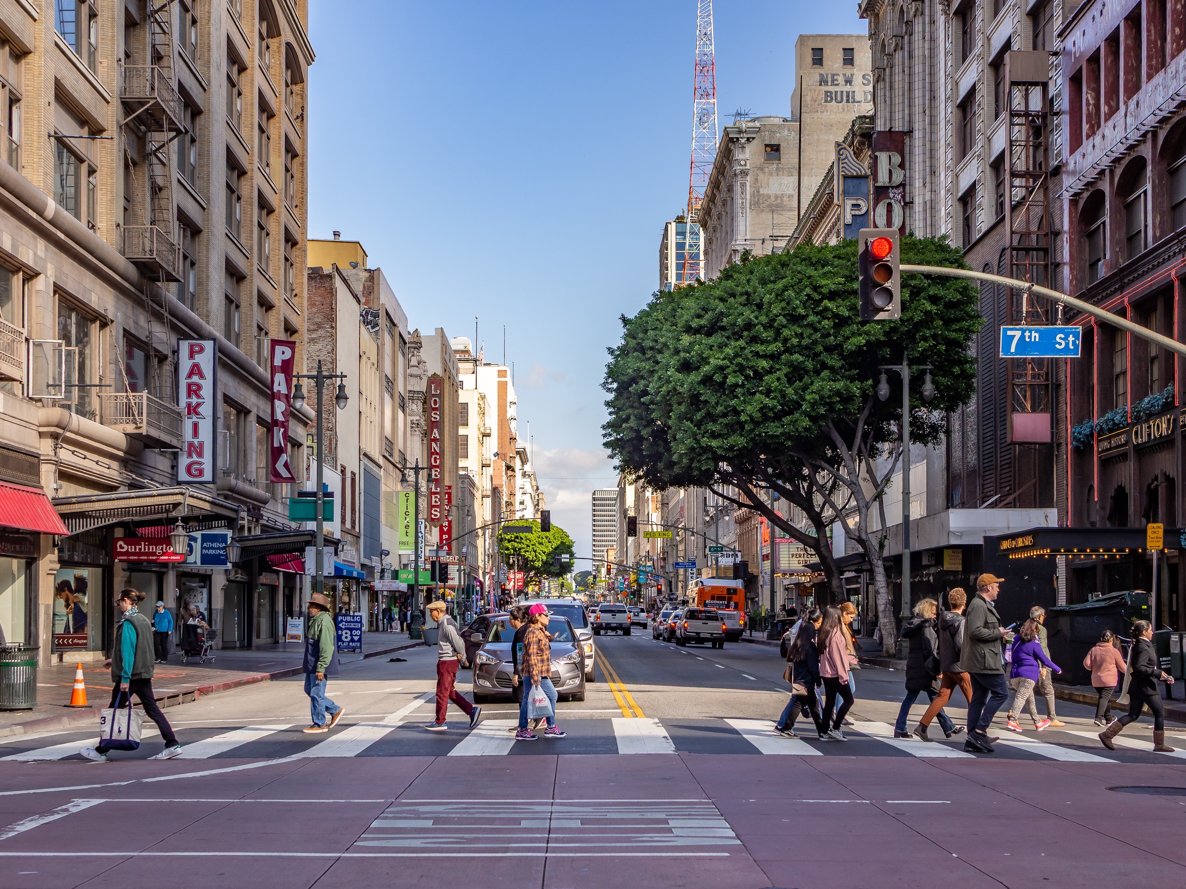 A busy street scene shows pedestrians crossing a road lined with tall buildings in downtown Los Angeles.