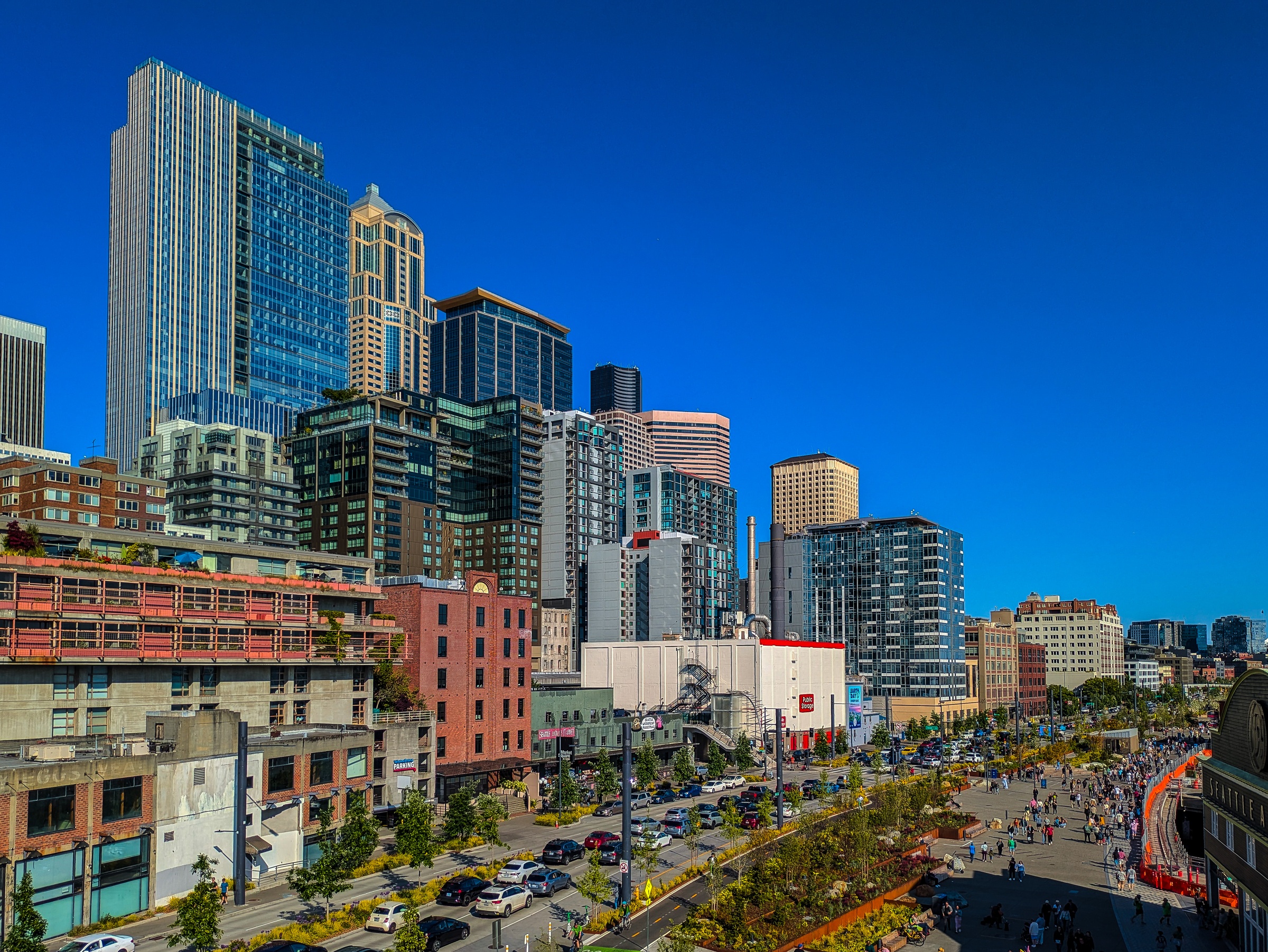 Seattle's skyline of modern skyscrapers rises above a waterfront promenade with pedestrians and trees.