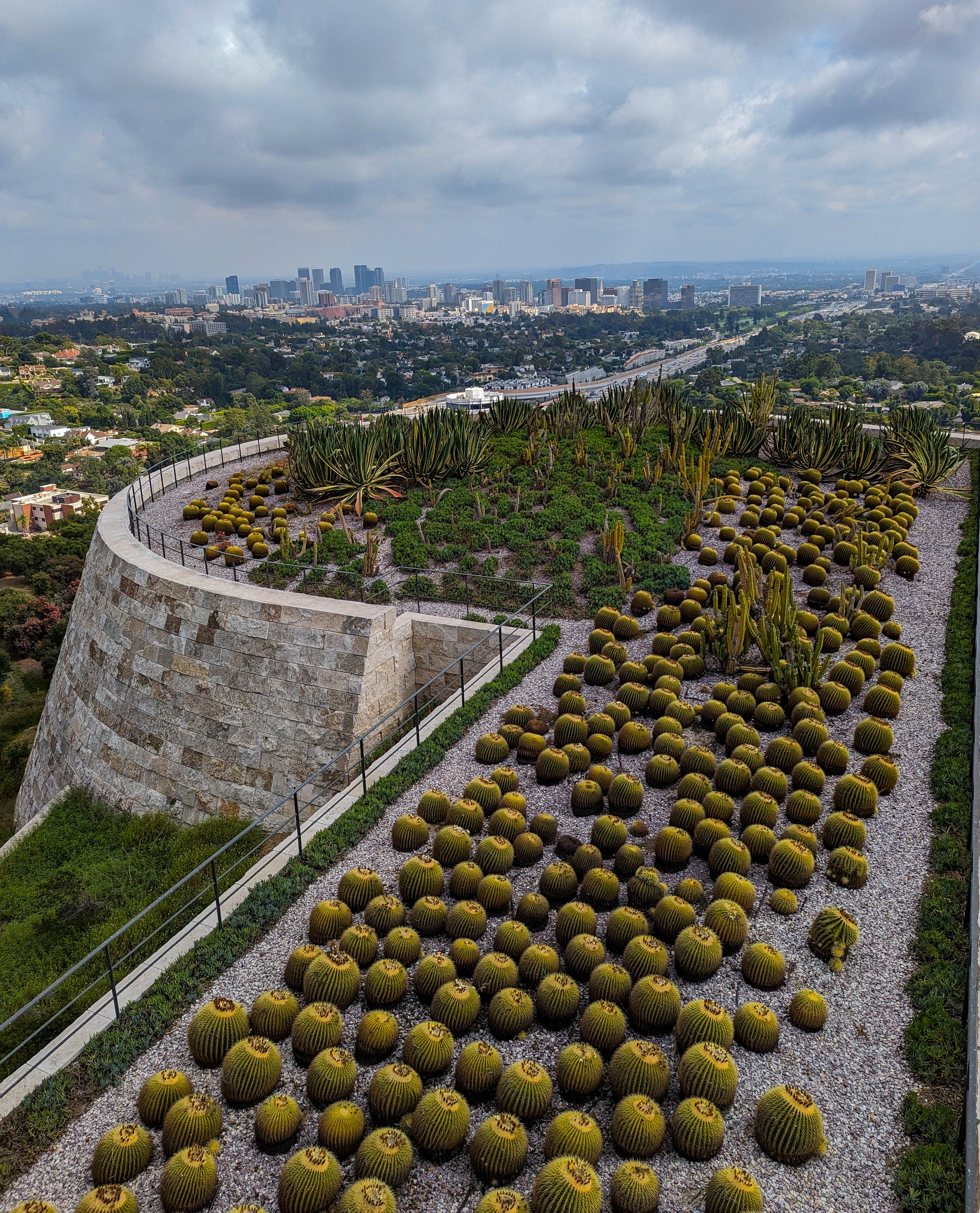 A stone structure with cacti and greenery overlooks a sprawling cityscape under an overcast sky.