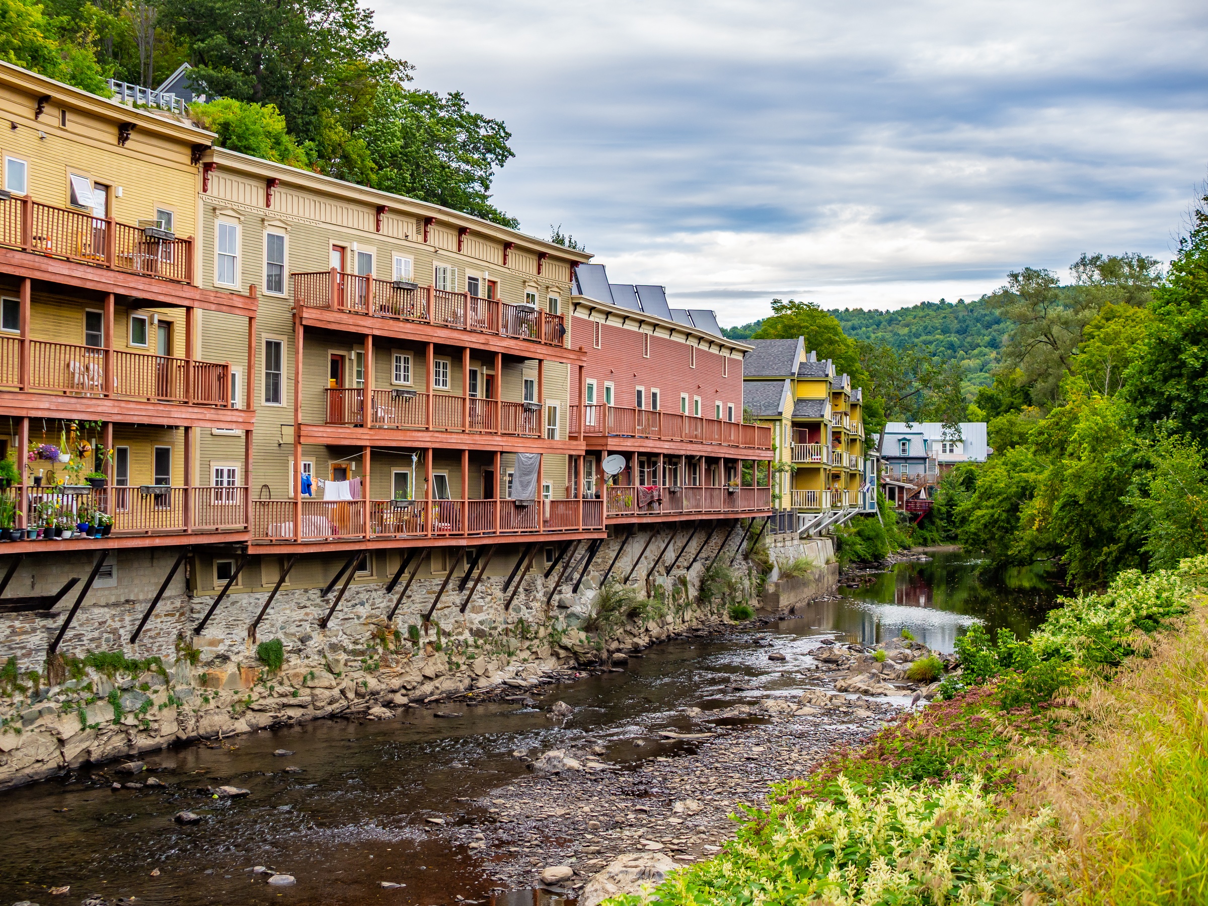 Colorful buildings with balconies line a riverbank, nestled within a lush green valley.