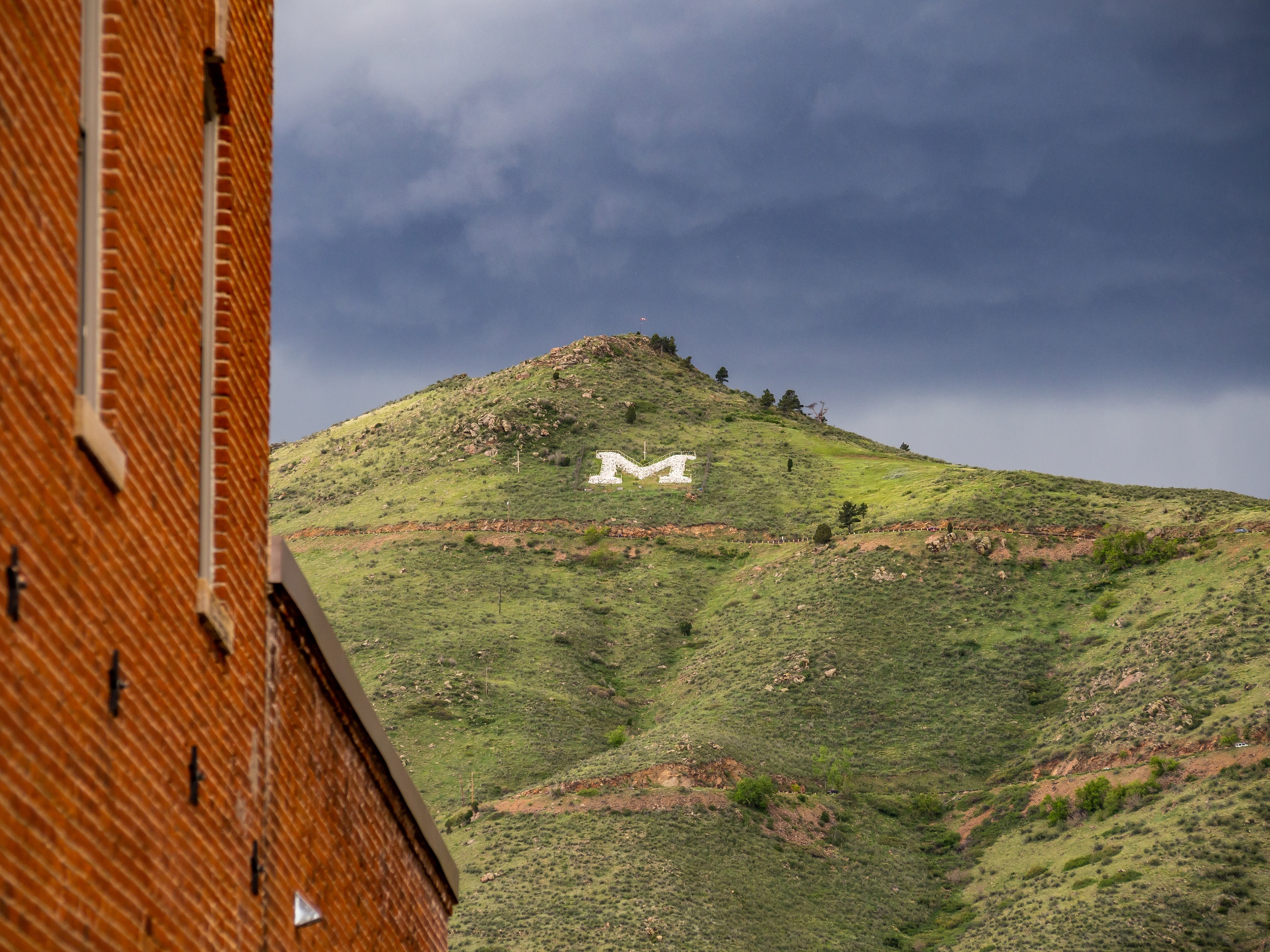 A large white "M" is displayed on a grassy hillside beneath a cloudy sky.