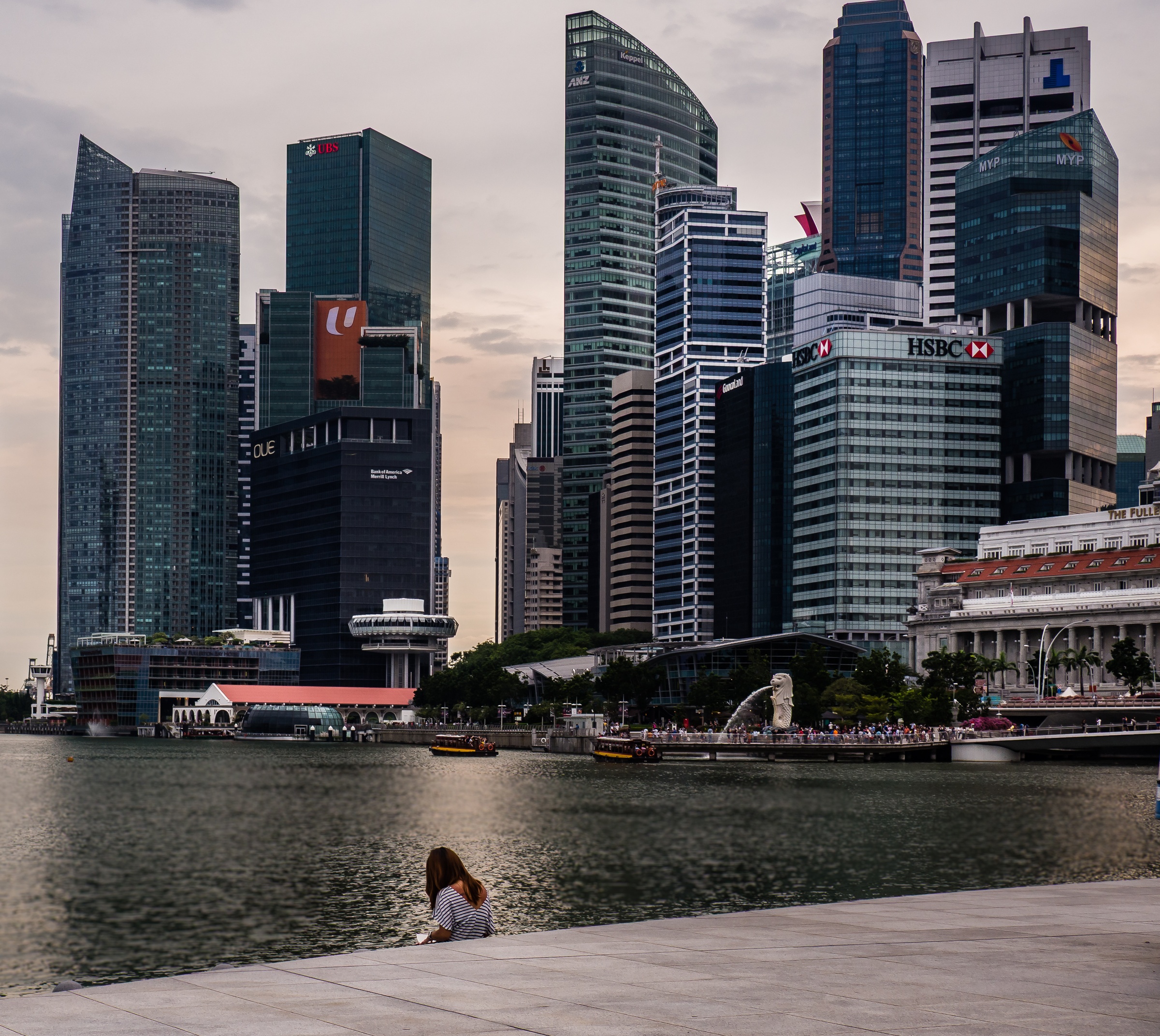 A person sits near a waterfront with modern skyscrapers rising in the background.