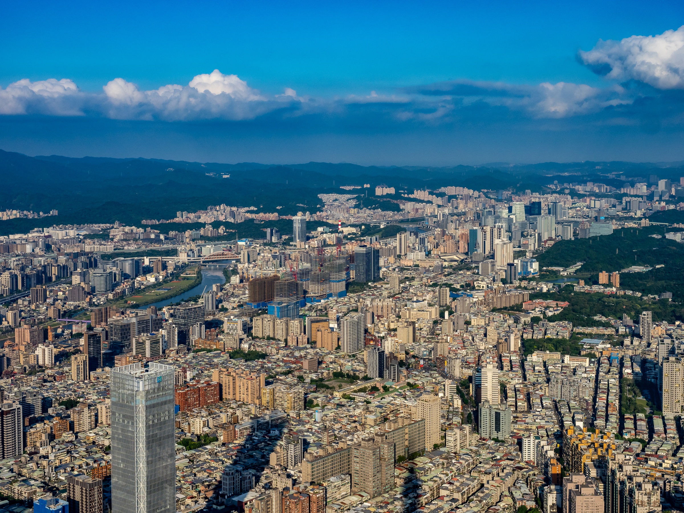 A sprawling cityscape with dense buildings, a river, and distant hills under a cloudy sky.