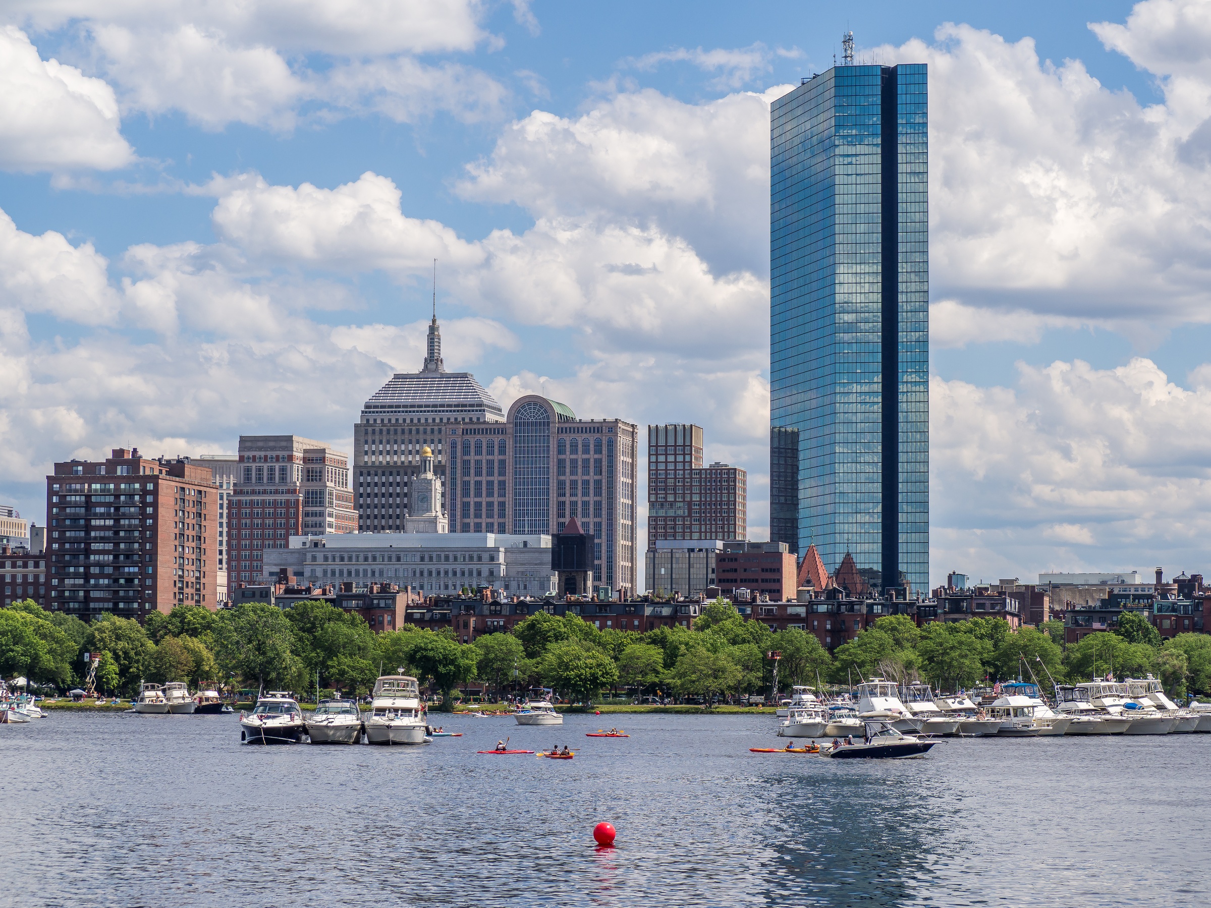 Boston's skyline rises above the Charles River with boats and a bright blue sky.