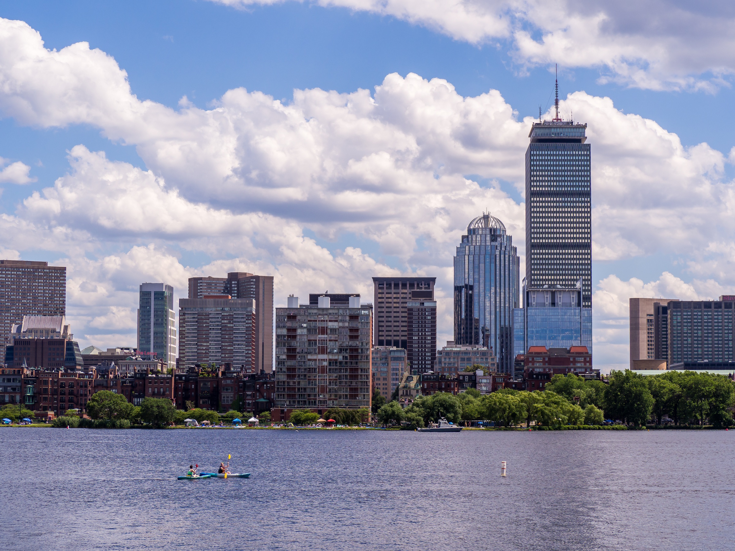 Boston's skyline rises above a calm river with people kayaking in the foreground.