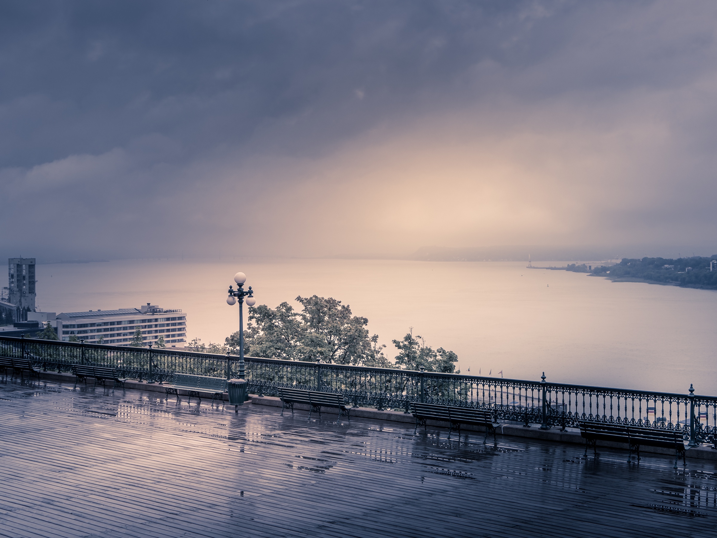 A wet promenade overlooks a body of water under an overcast sky with distant buildings.