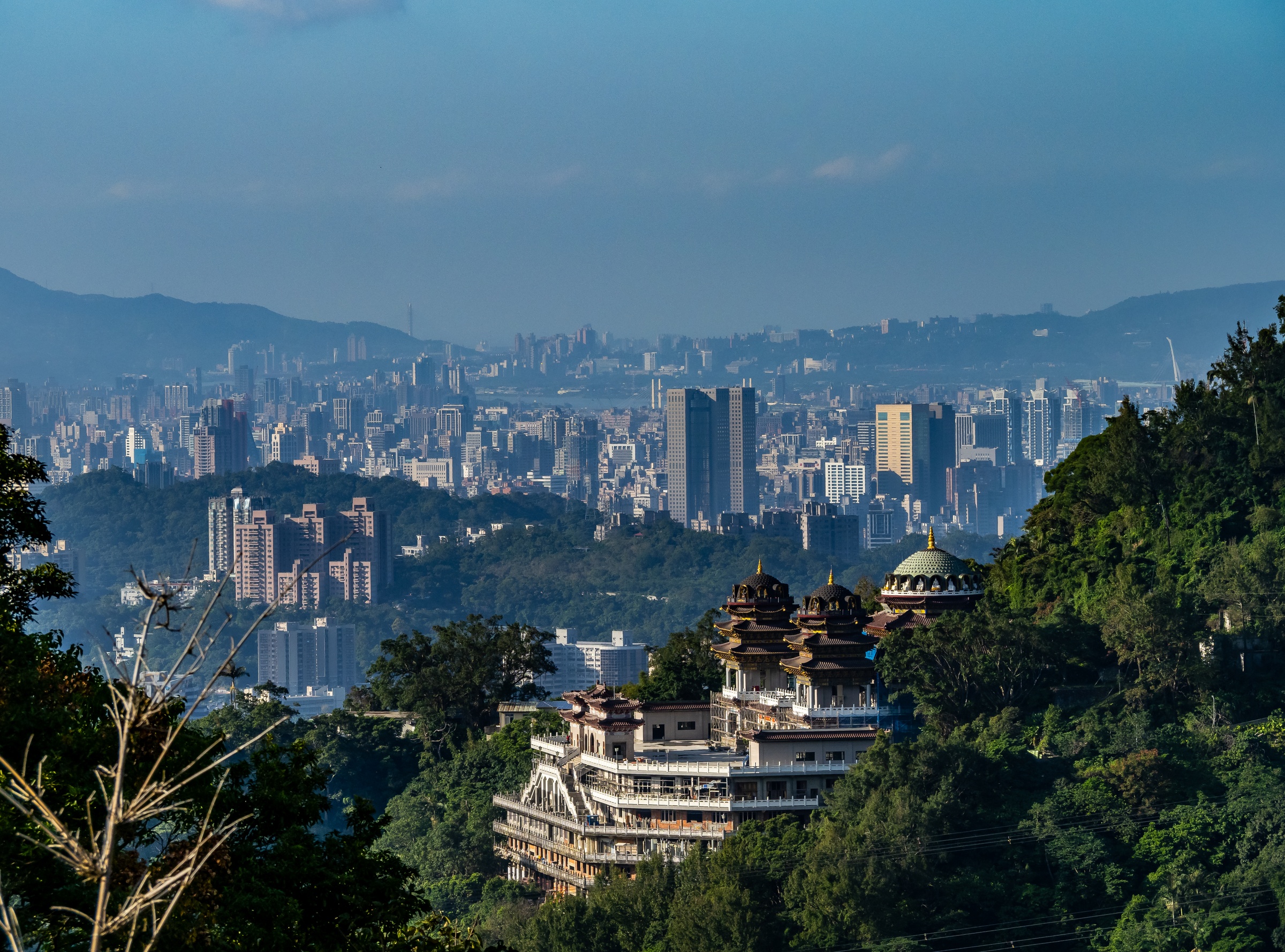 A temple complex sits atop a green hill overlooking a sprawling cityscape in the distance.