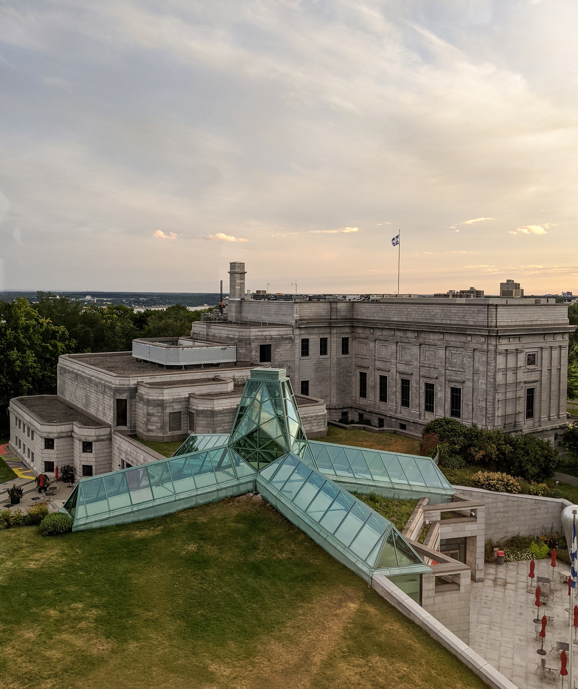 A modern glass structure extends from a stone building overlooking green space and a distant cityscape.