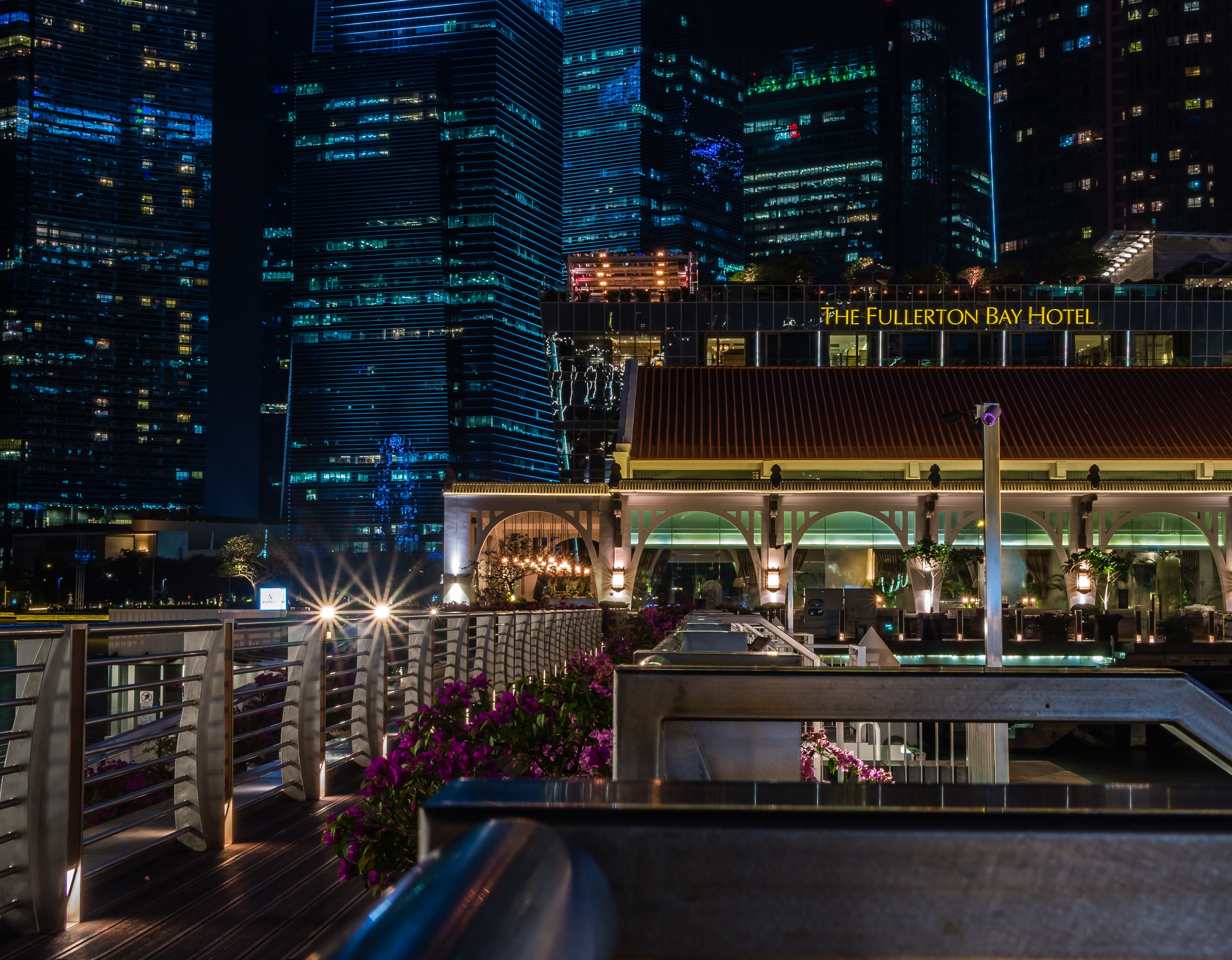The Fullerton Bay Hotel sits below illuminated skyscrapers along a waterfront promenade at night.