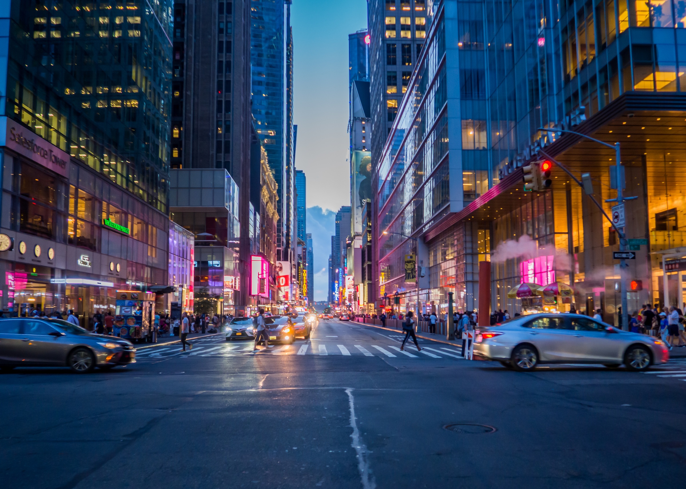 Cars and pedestrians traverse a busy, brightly lit street canyon in midtown Manhattan at dusk.