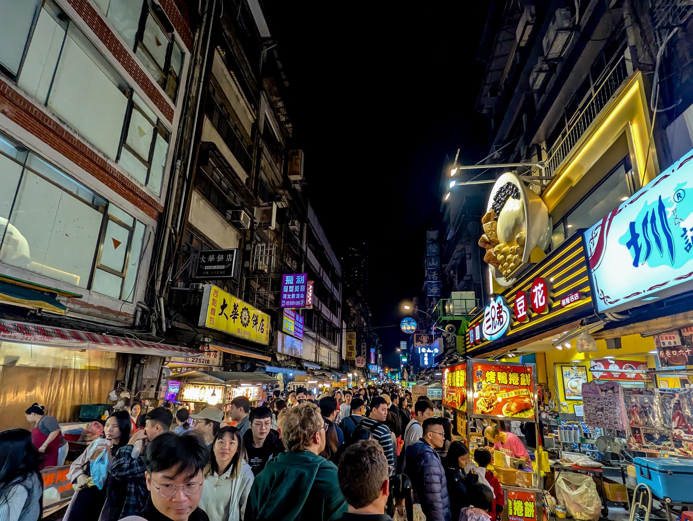A crowded night market fills a narrow street lined with tall buildings and illuminated signs.