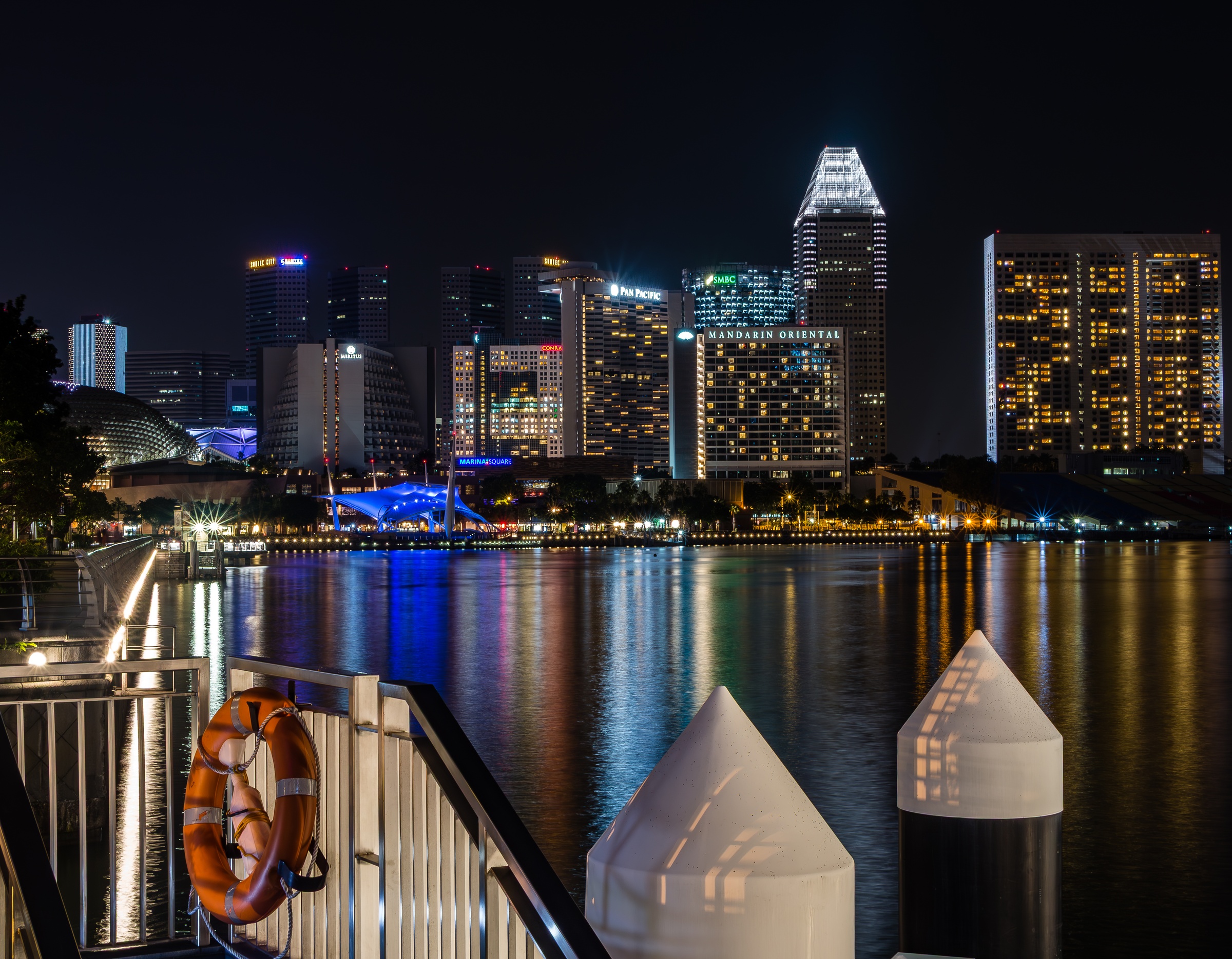 Singapore's illuminated skyline reflects in the water, framed by a walkway and nautical markers.