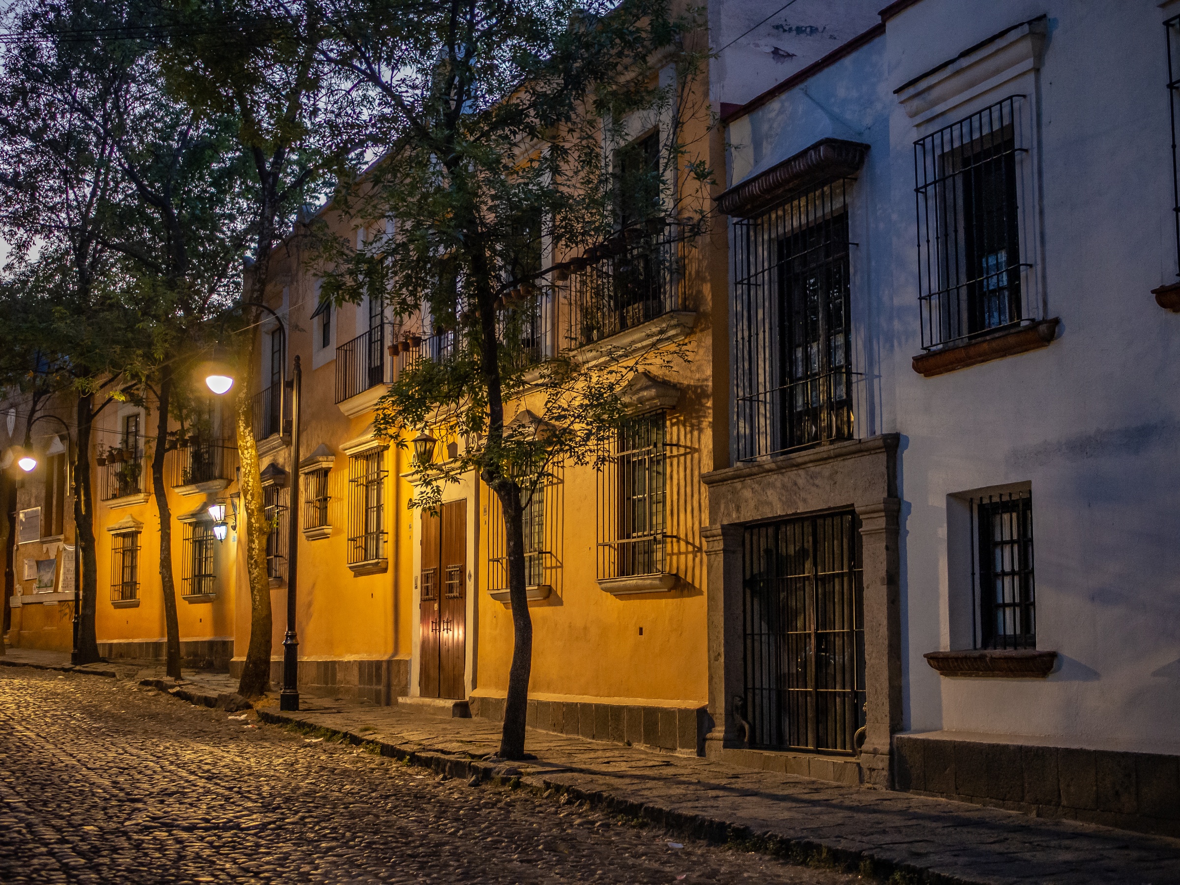 A row of yellow buildings with wrought-iron balconies lines a cobblestone street at dusk.