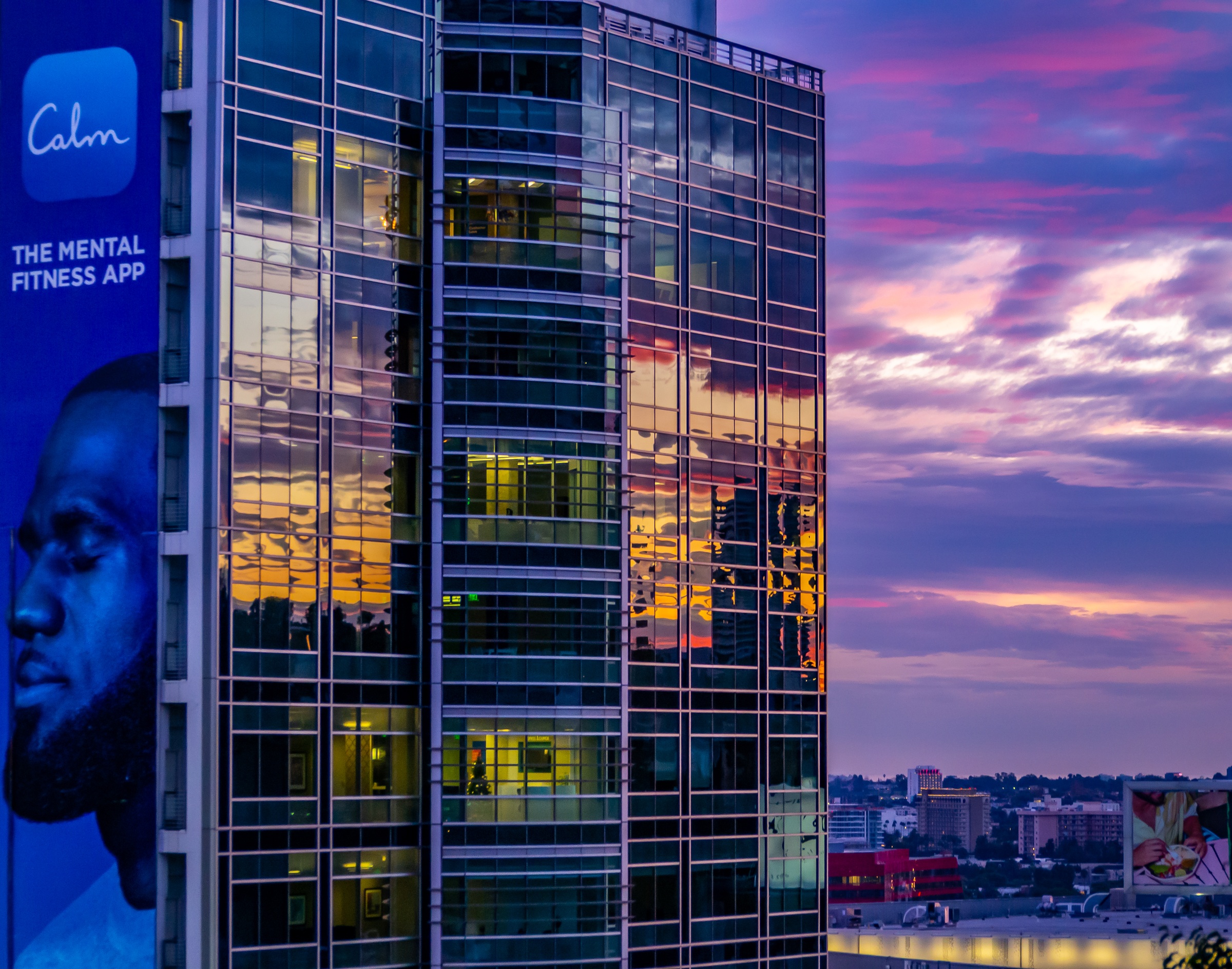 A modern glass building reflects a sunset sky, with a large advertisement on its side.