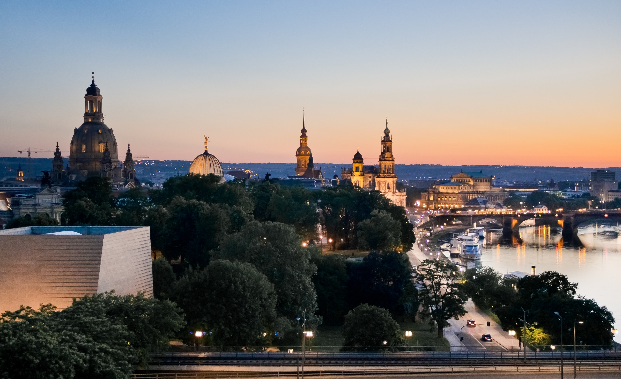 Dresden's skyline with historic buildings and a river glows at dusk under an orange sky.