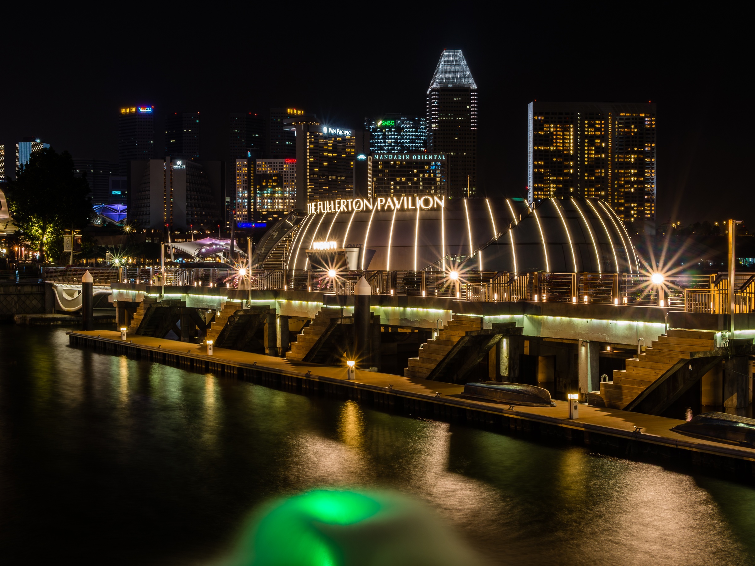 The Fullerton Pavilion sits on a waterfront promenade with Singapore's skyline illuminated in the background.