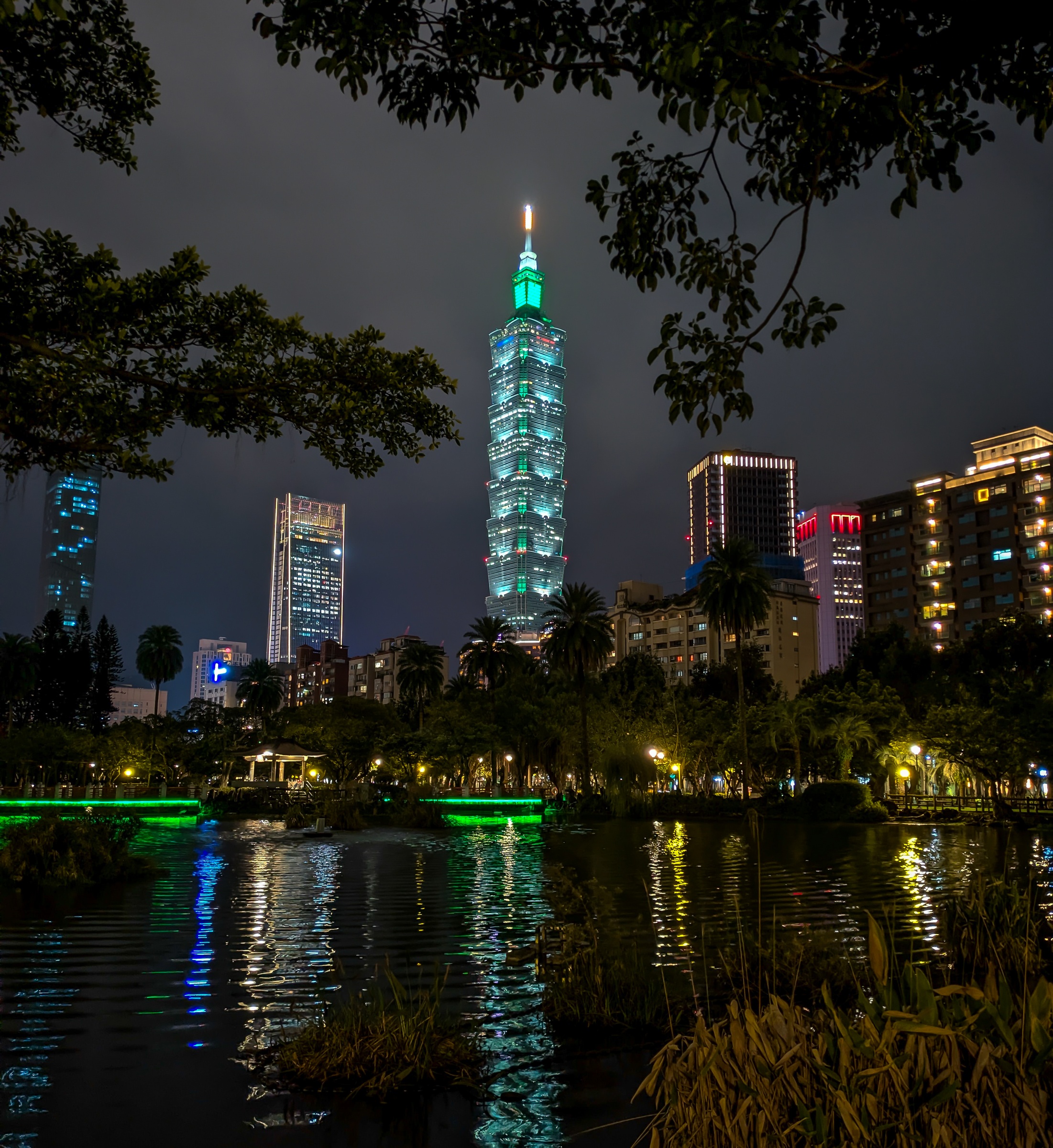 Taipei 101 tower glows above a pond and park with other buildings in the night.
