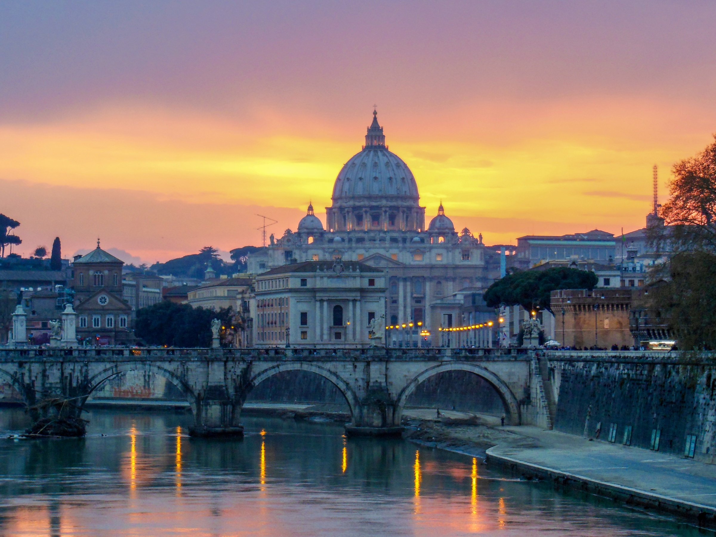 St. Peter's Basilica and surrounding buildings are silhouetted against a vibrant sunset over the Tiber River.