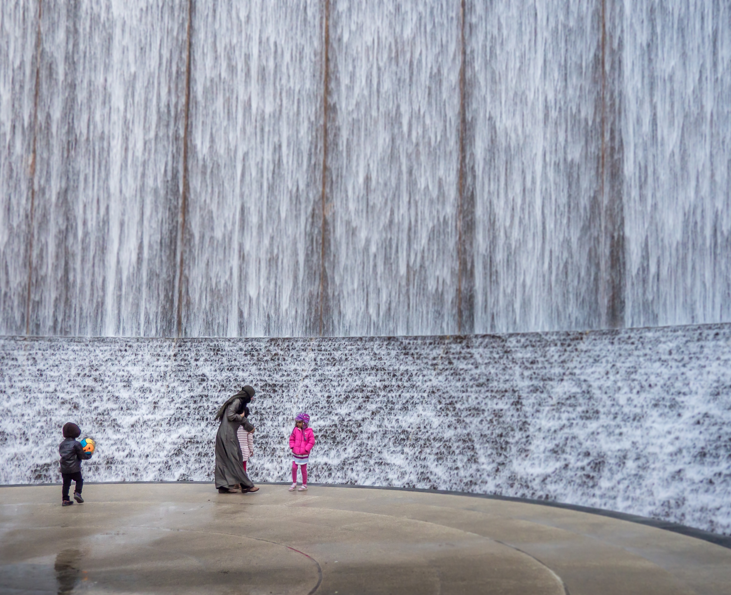 A woman with a child stands near two other children in front of a large water curtain.