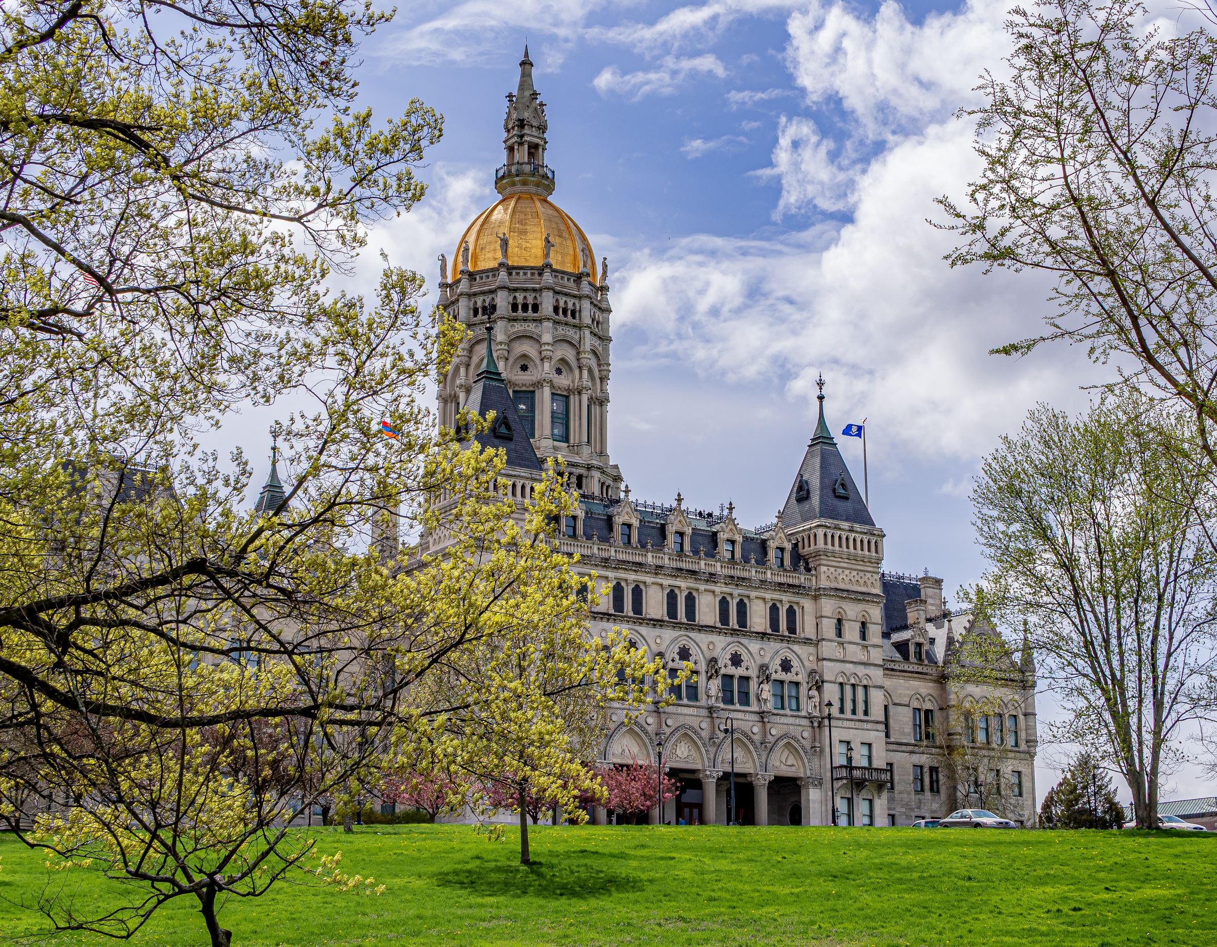 The gold-domed Connecticut State Capitol building stands framed by blossoming trees and a green lawn.