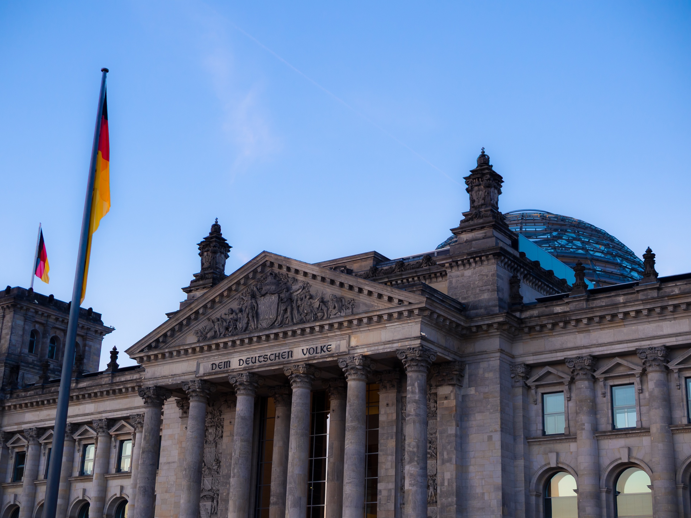 The German flag flies over the Reichstag building in Berlin against a clear blue sky.