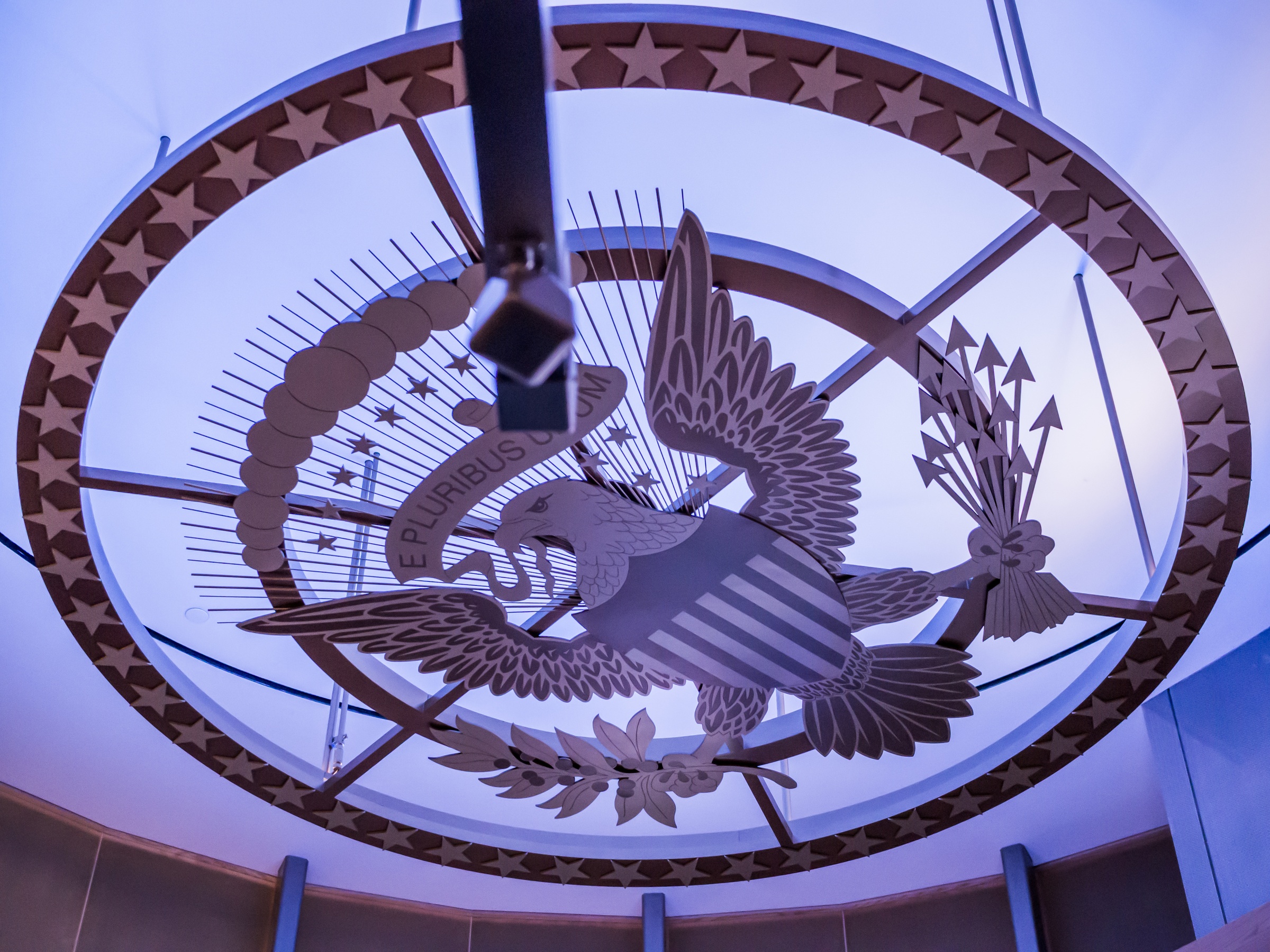 A large, ornate eagle and shield sculpture hangs from a ceiling within a building.