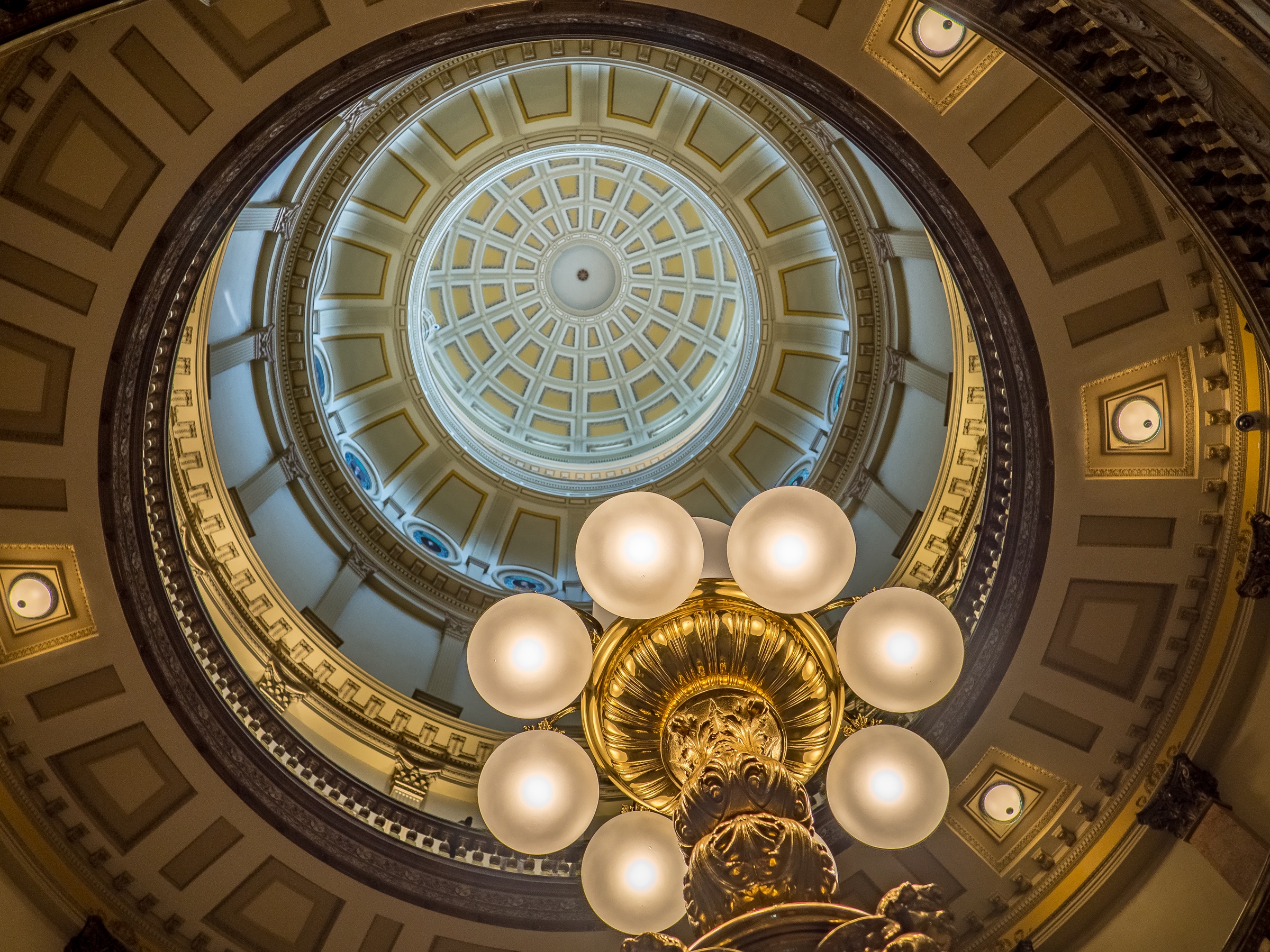 An ornate chandelier hangs beneath a grand, domed ceiling with decorative panels and recessed lighting.