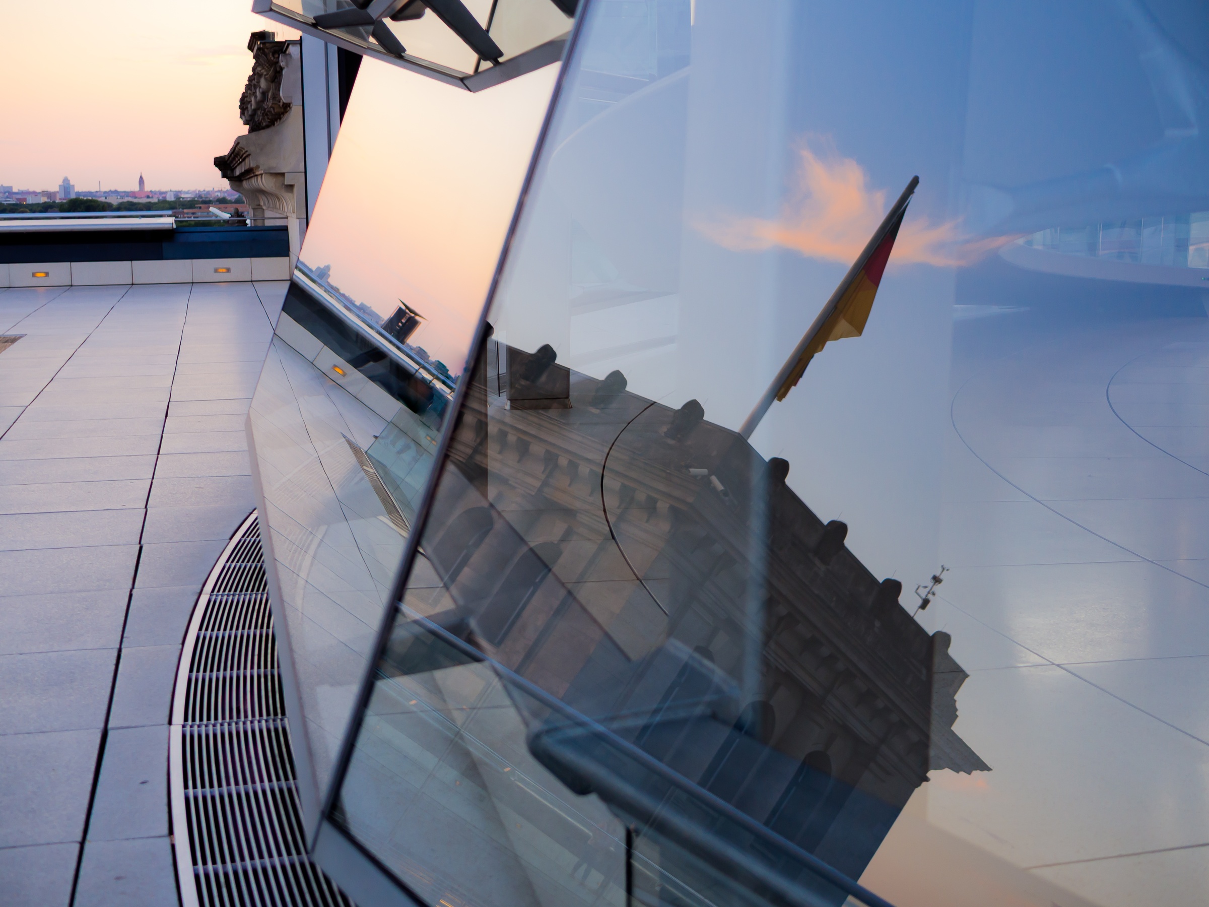 A German flag reflects in a large glass window overlooking a cityscape at dusk.