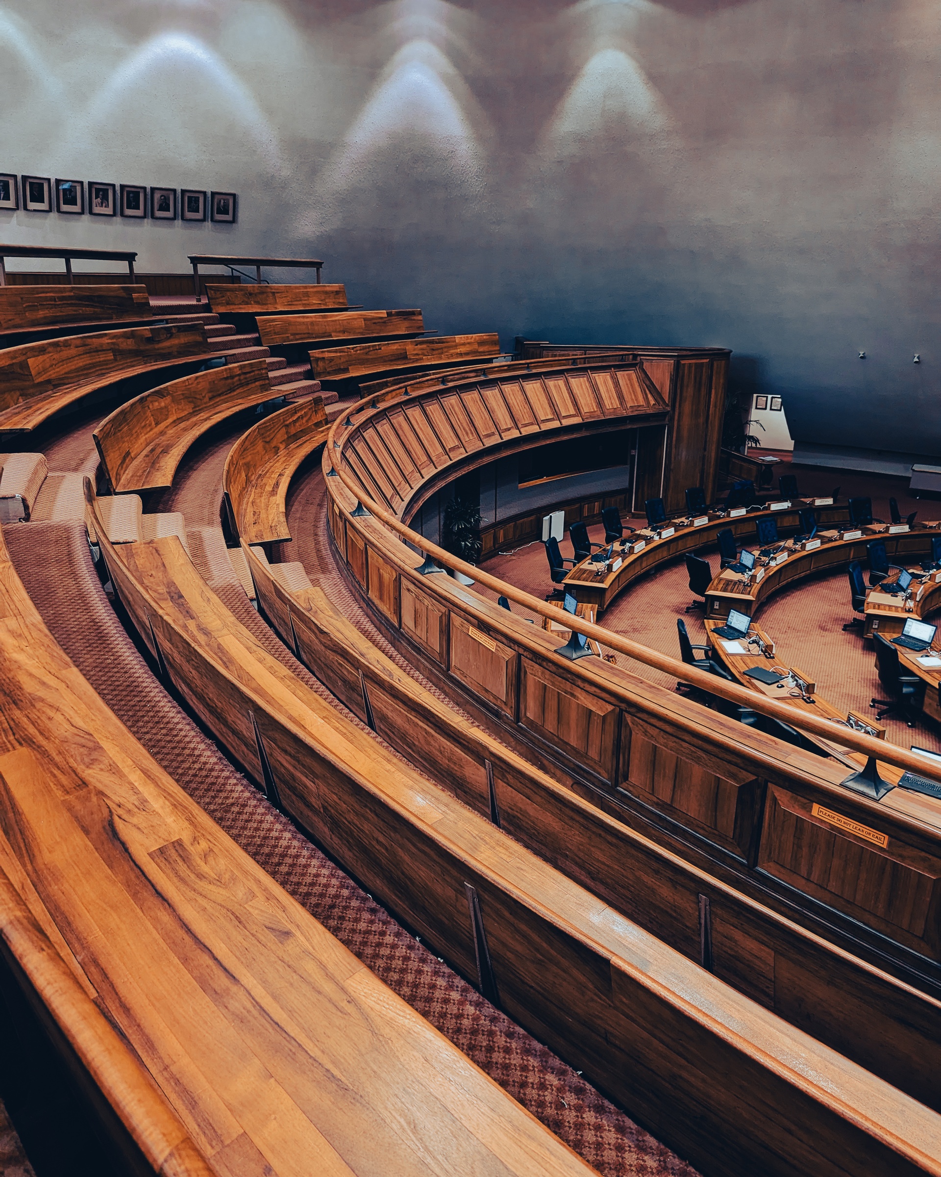 An auditorium with tiered wooden seating curves around a central area featuring desks and a raised platform.