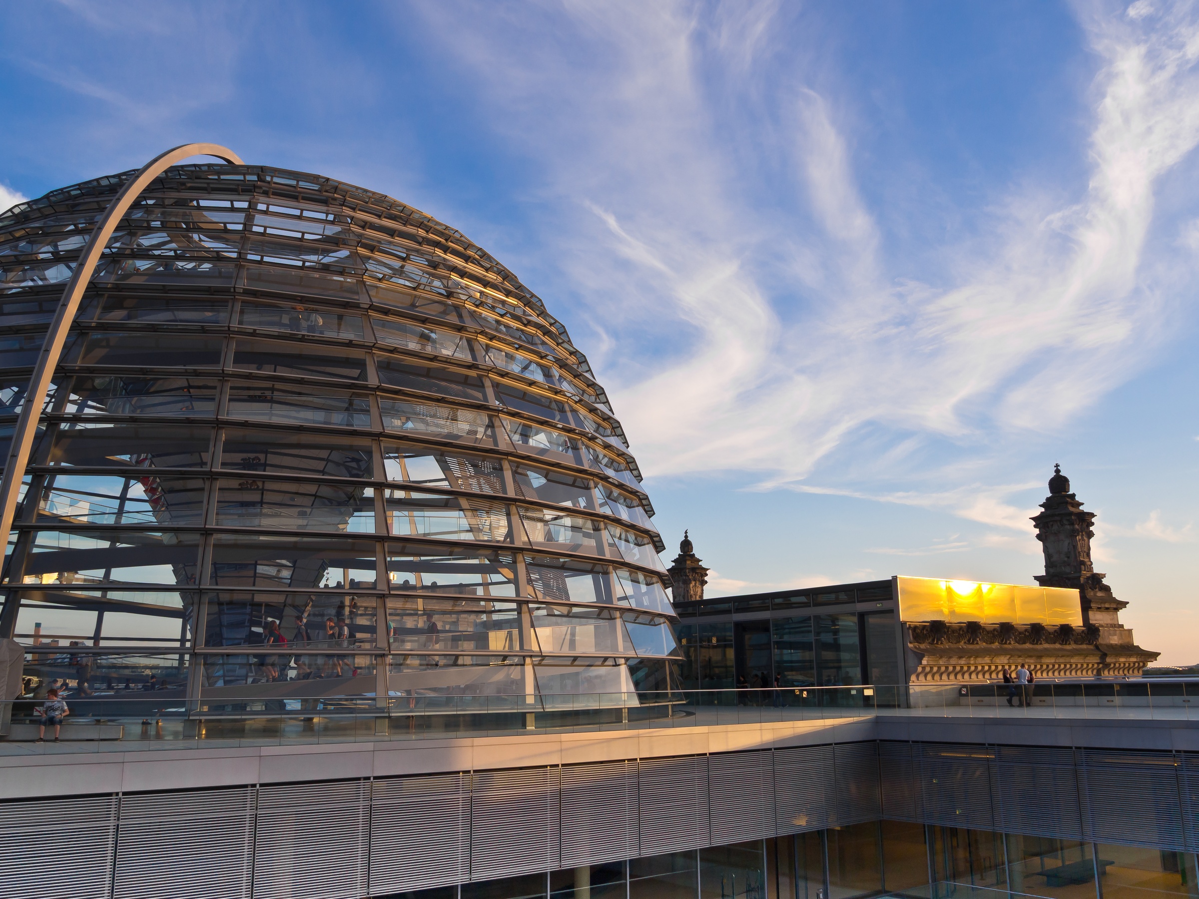 The glass dome of the Reichstag building overlooks a monument against a partly cloudy sky.