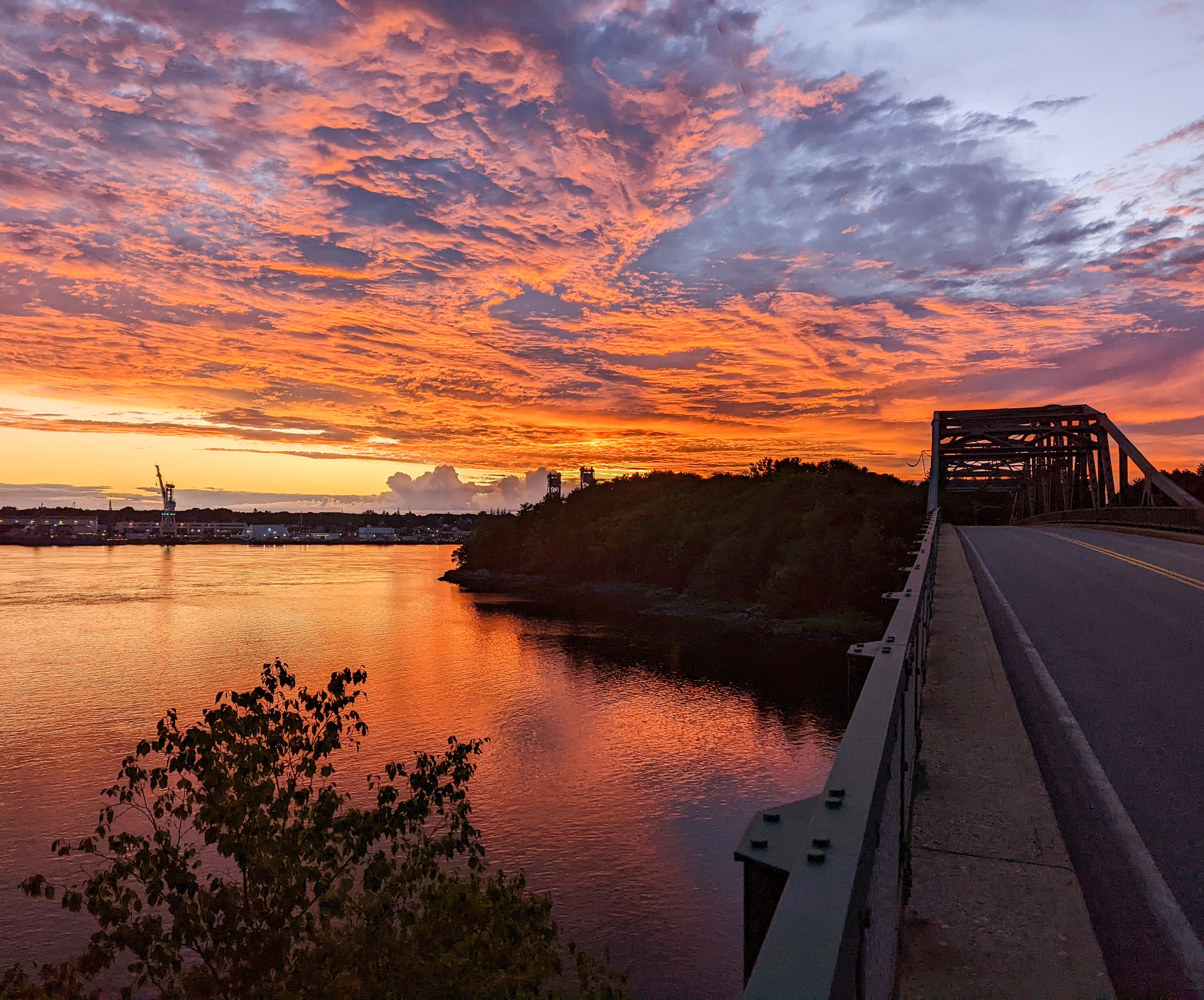 A vibrant sunset illuminates a bridge over water with trees and distant cranes visible.