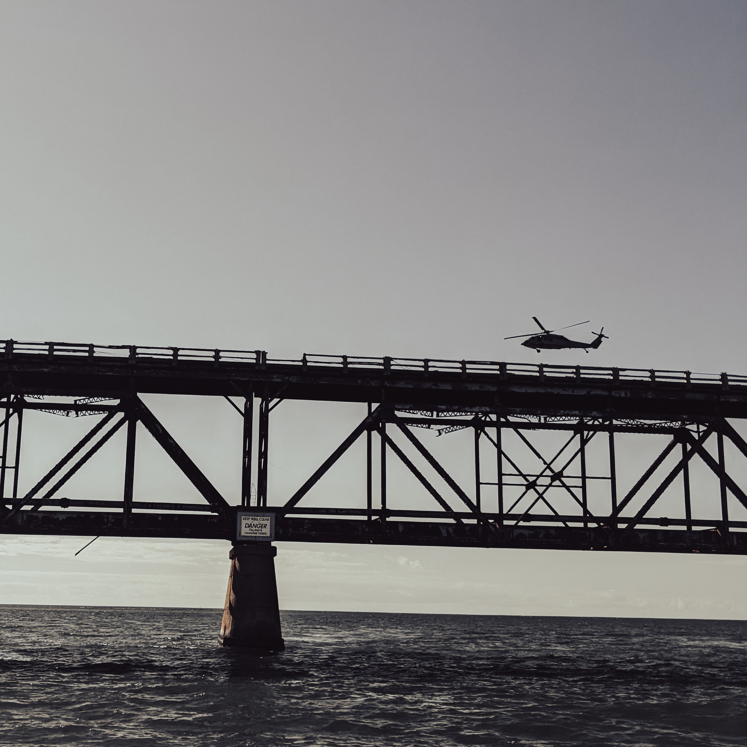 A helicopter flies above a steel bridge extending over dark water under a clear sky.