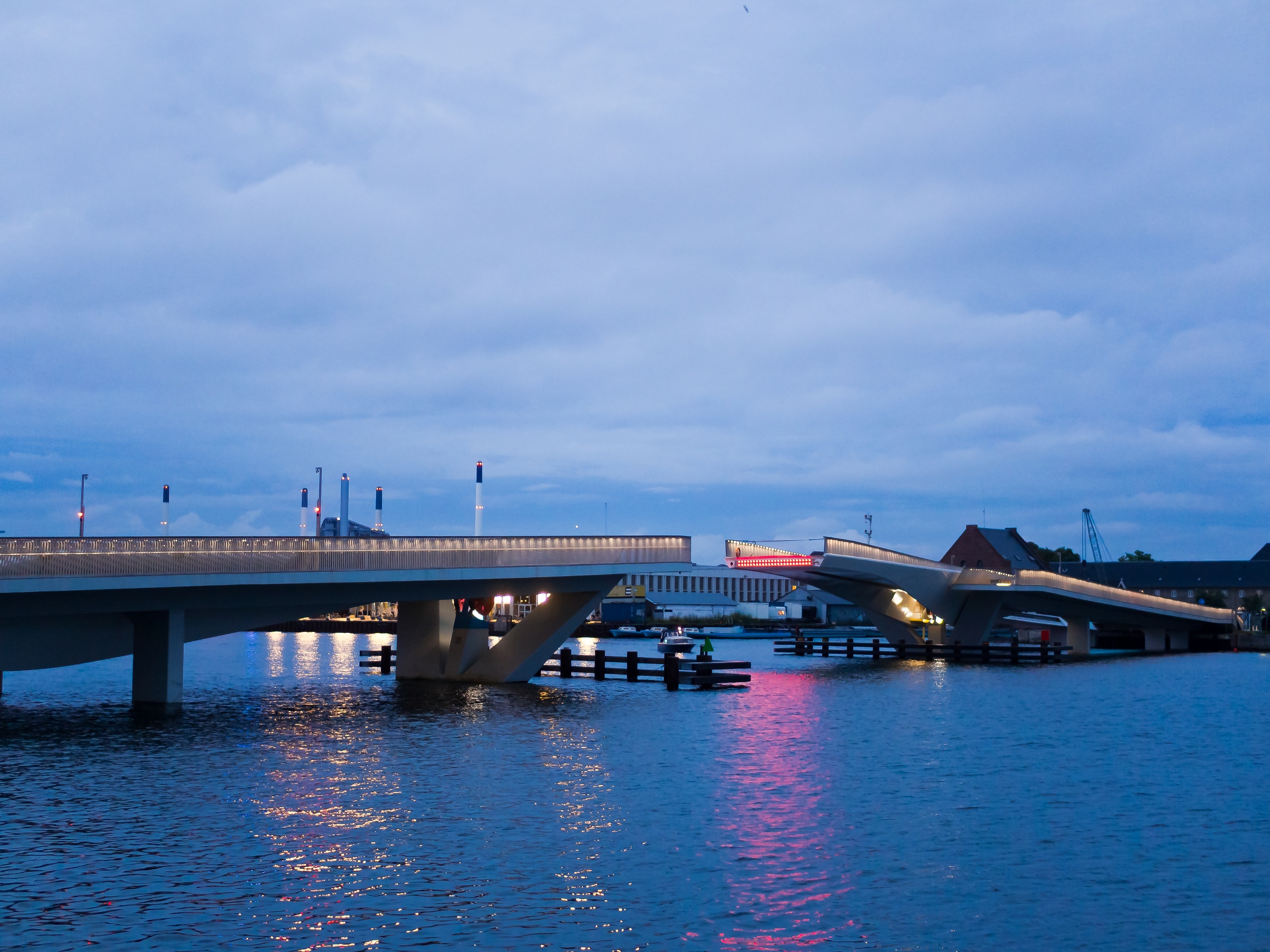 A bridge with illuminated sections spans a body of water under an overcast twilight sky.