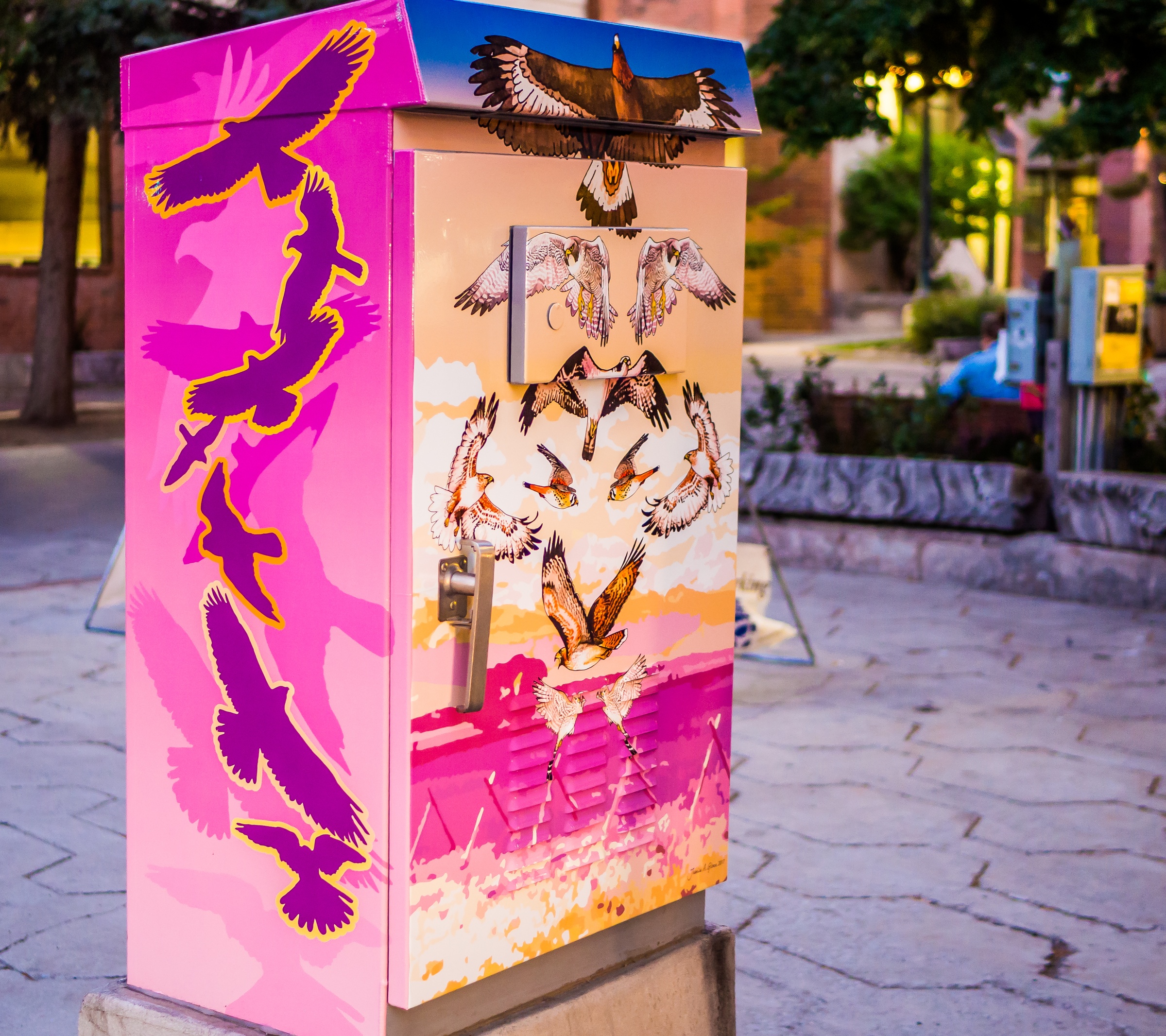 A painted utility box depicts birds against a pink and orange sunset in an urban plaza.
