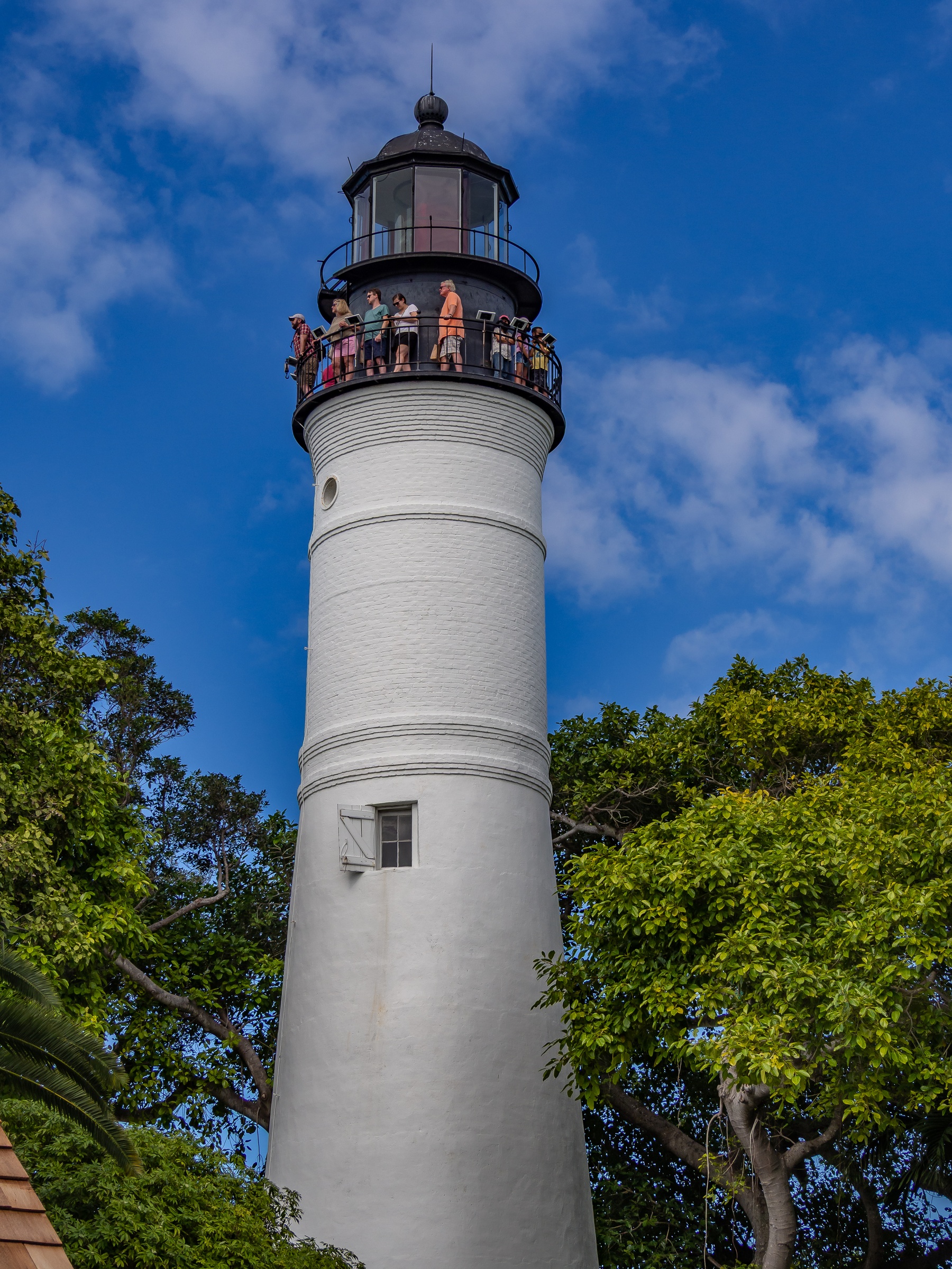 A white lighthouse stands tall against a blue sky, framed by lush green trees.