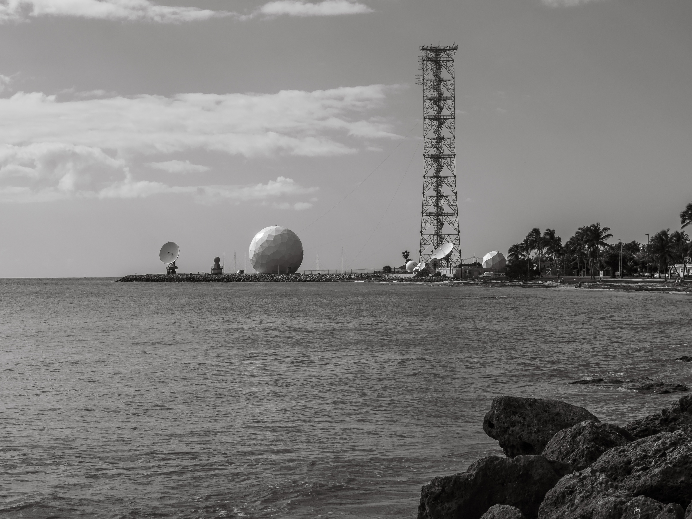 A tall tower and satellite dishes stand along a shoreline with large spherical sculptures overlooking the ocean.